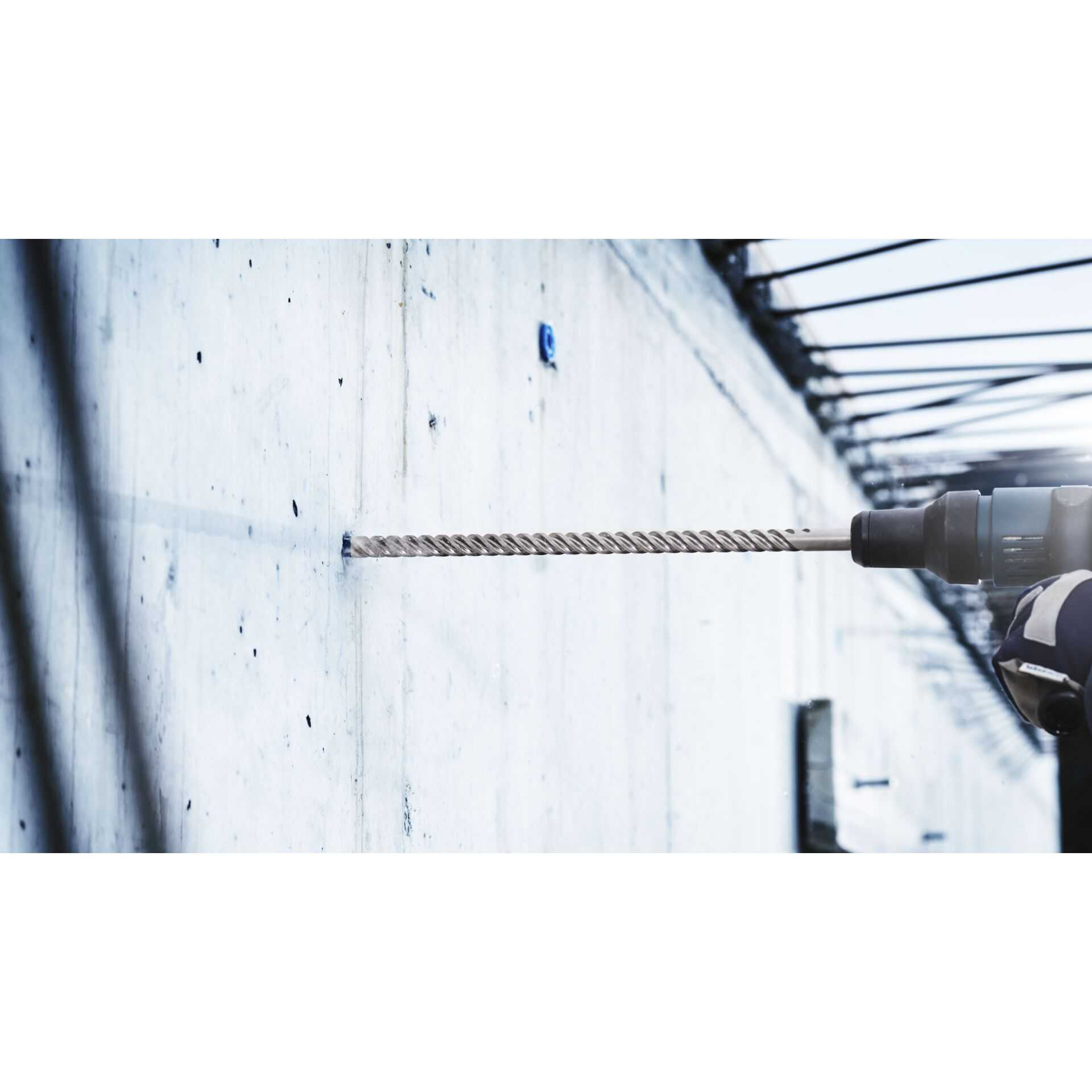 Wiertło wiercące w betonowej ścianie. Widoczny pył i gruz. Jasnoniebieskie tło ściany.