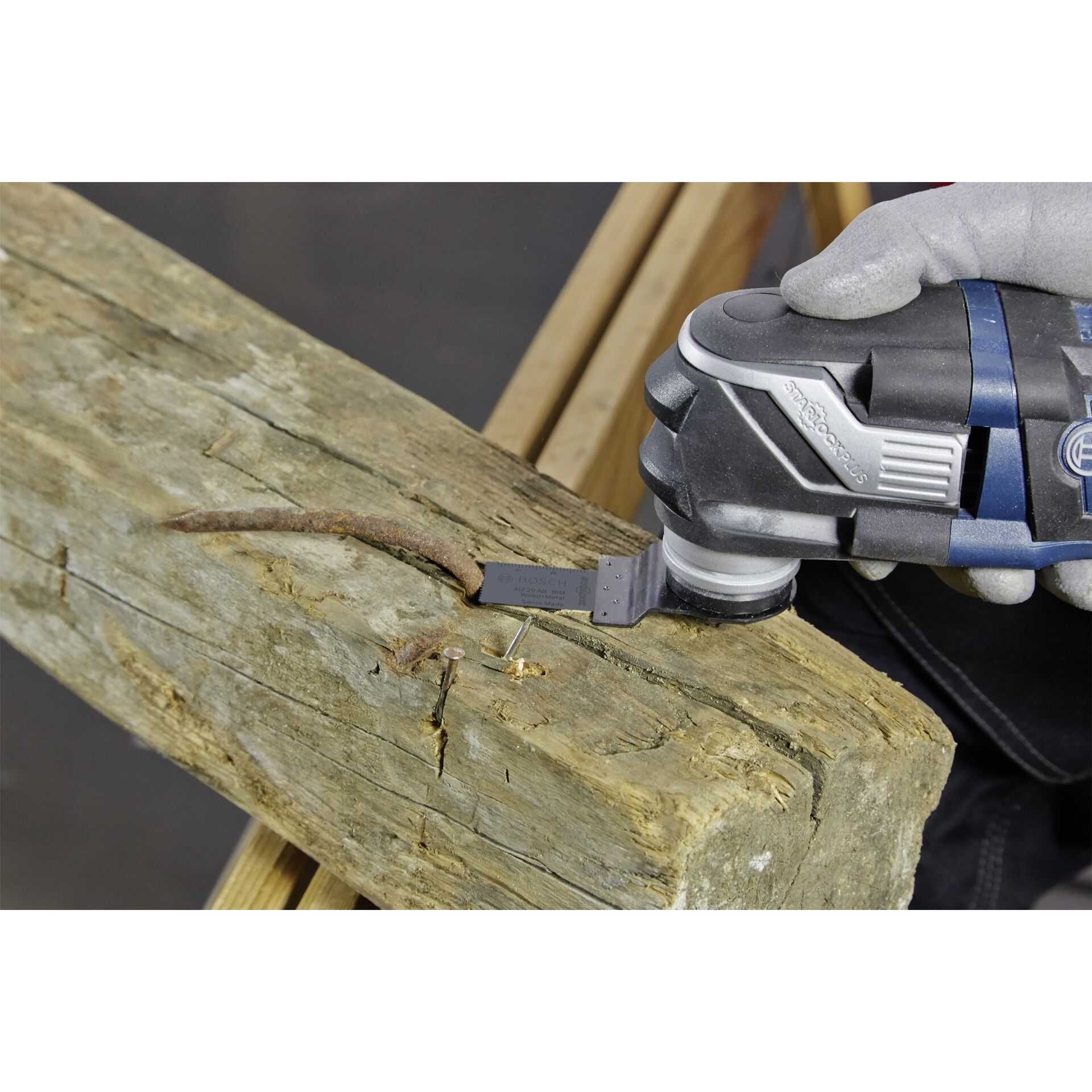A multi-tool cutting a nail in a wooden beam. The tool is held by a gloved hand.