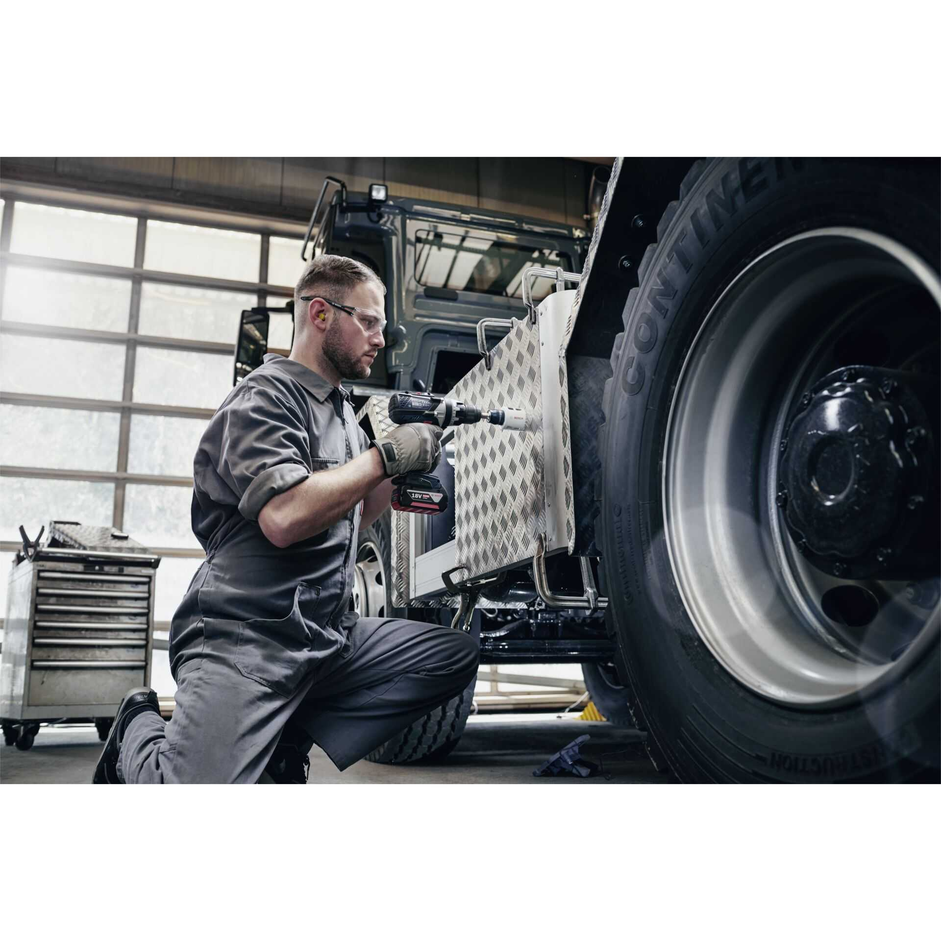 Mężczyzna używa wiertarki w pojeździe. Ma na sobie odzież roboczą i okulary ochronne.