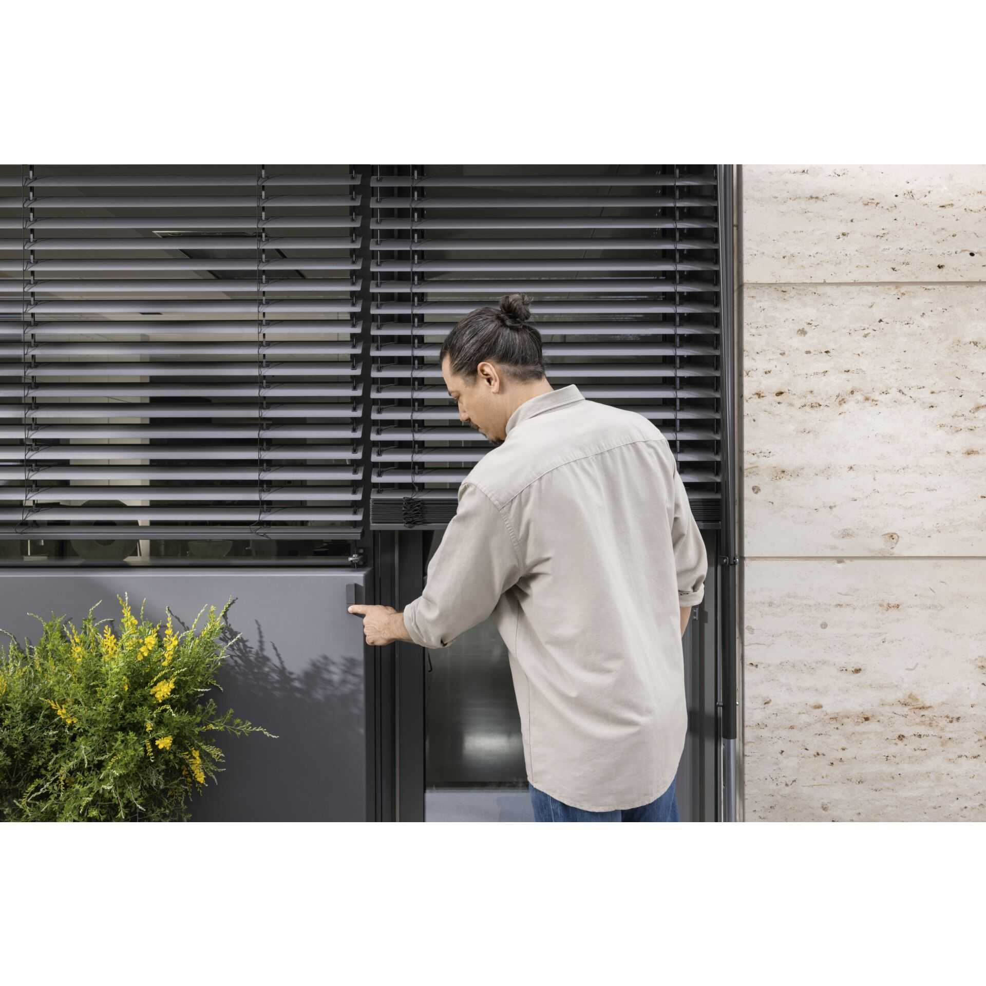 Mężczyzna regulujący żaluzje. Okno i kilka żółtych kwiatów w tle.