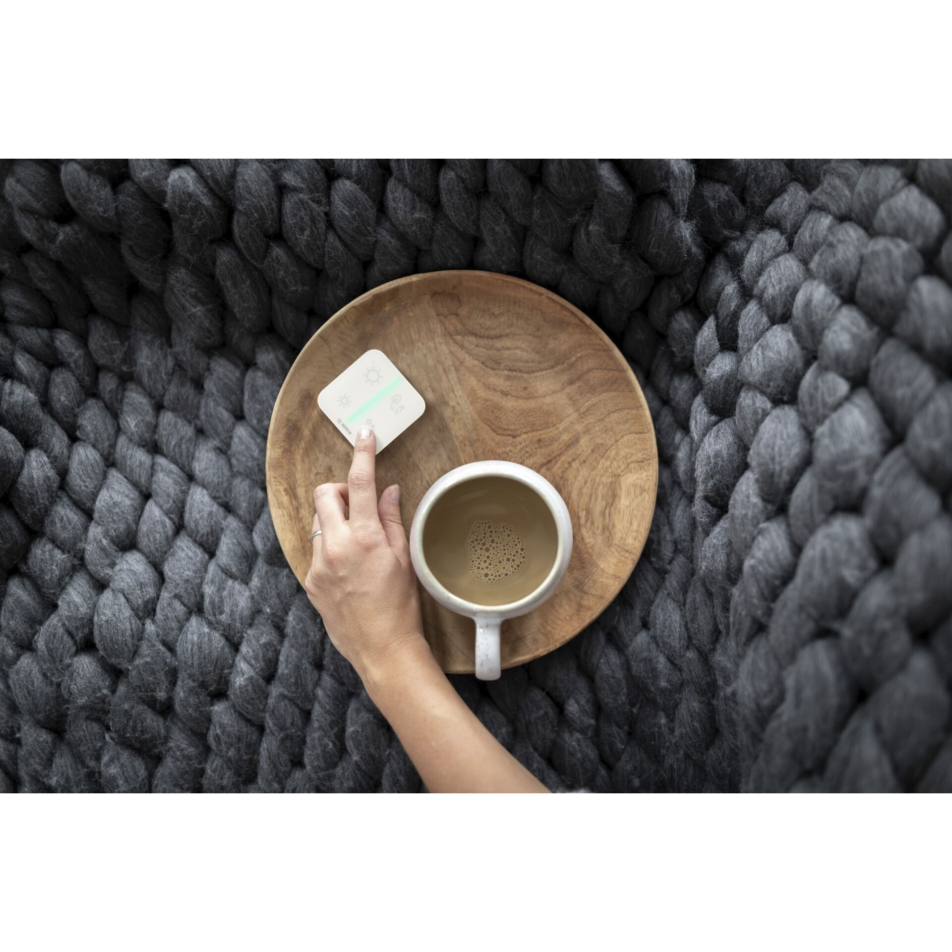 Eine Hand greift nach einem Smart-Home-Bedienfeld. Ein Holztablett mit einer Tasse Kaffee steht daneben.