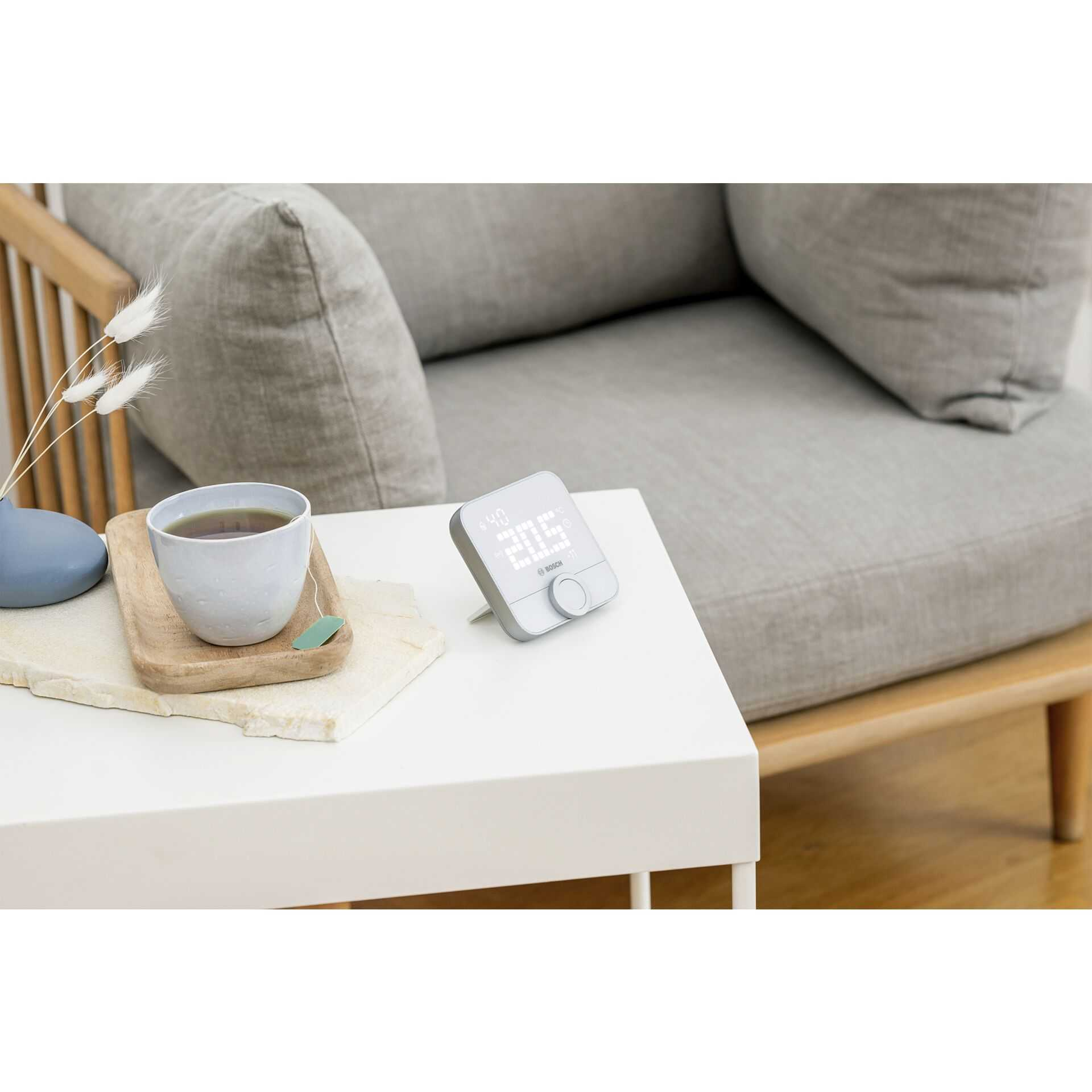 Ein Smart-Thermostat auf einem weißen Tisch. Eine Tasse Tee und ein Stuhl im Hintergrund.