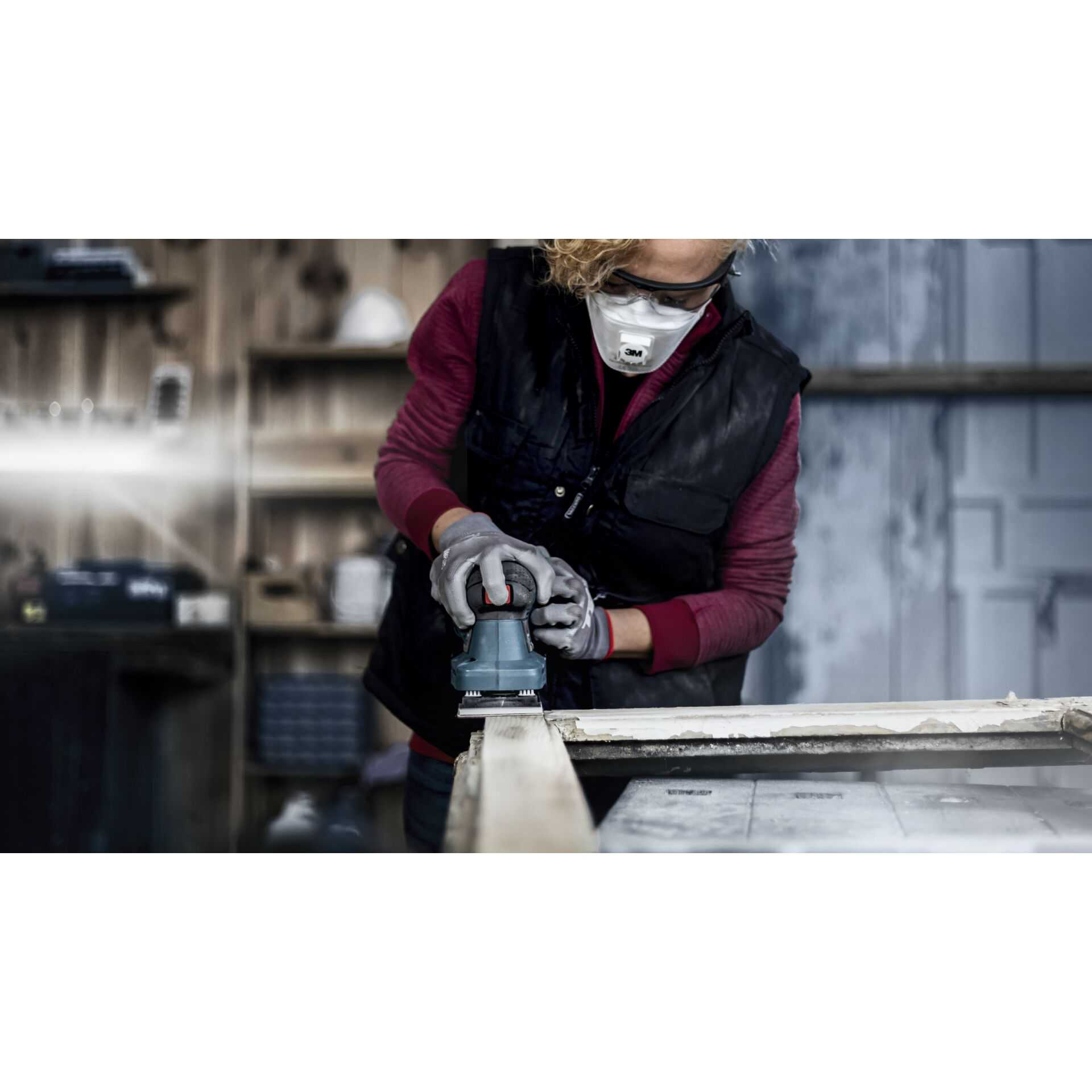 Persona che leviga un telaio in legno, indossa una maschera e guanti, in un'officina.