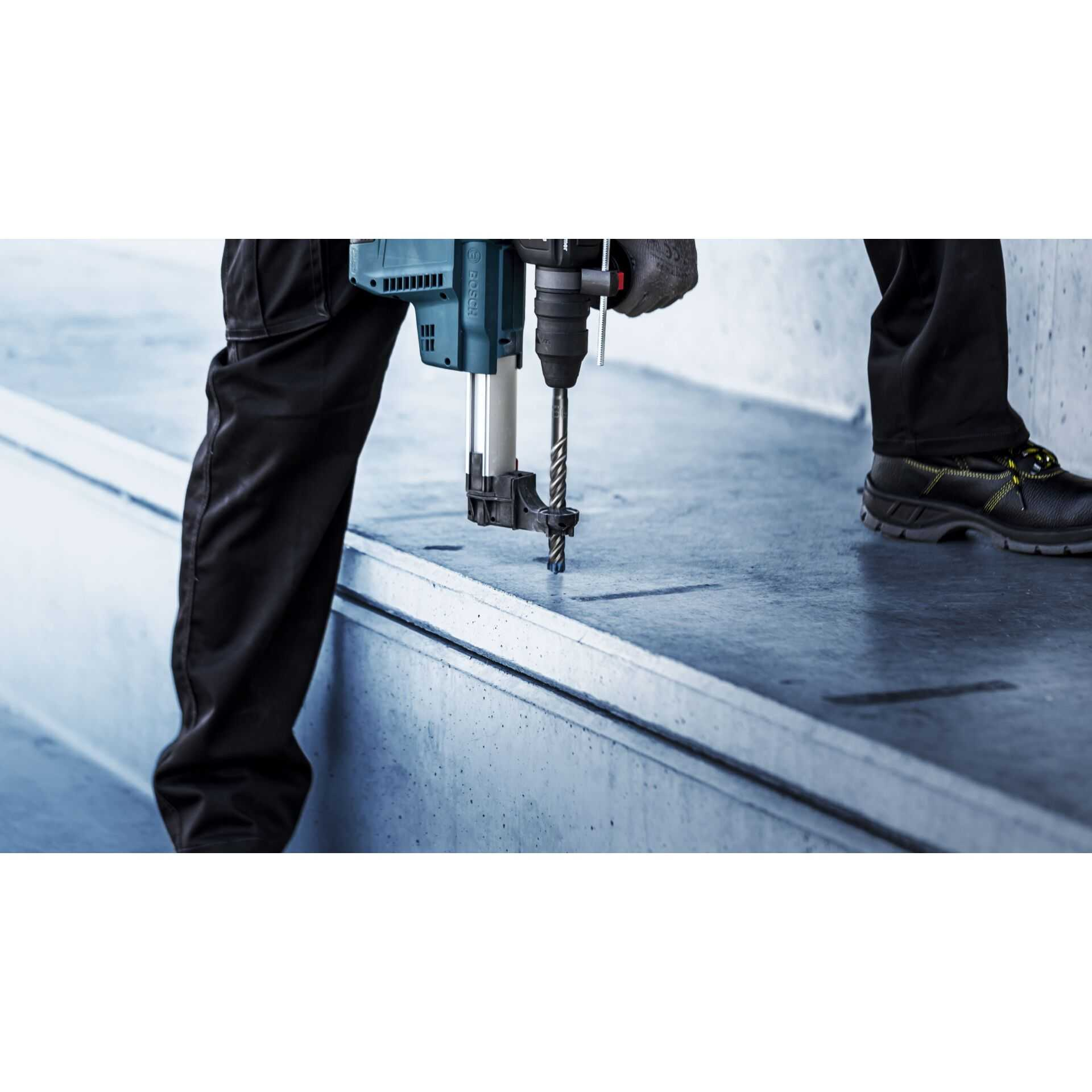 Osoba wiercąca w betonie niebieską wiertarką Bosch. Widoczne czarne spodnie i buty. Szare betonowe tło.