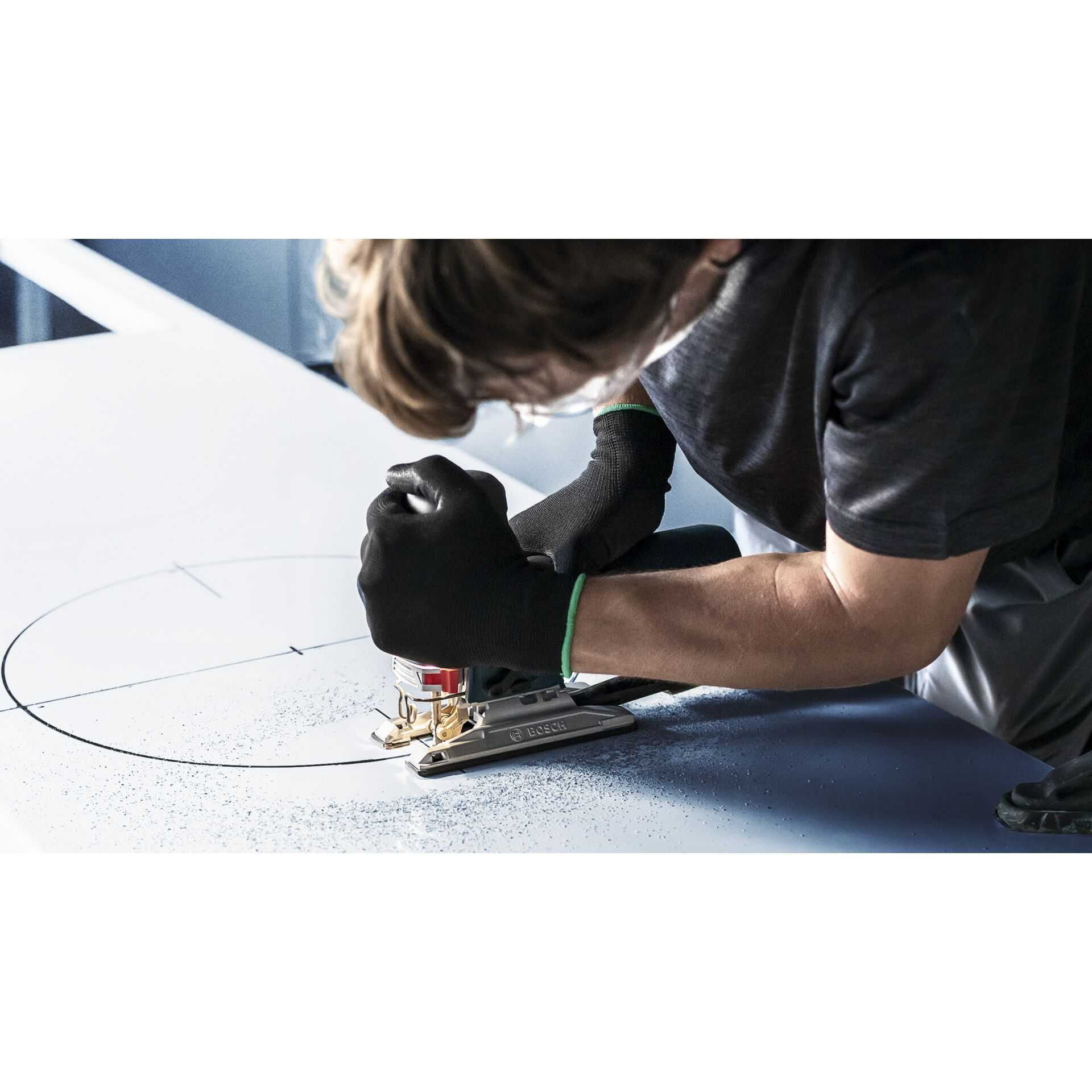 Osoba wycina koło w białym materiale za pomocą wyrzynarki Bosch. Czarne rękawice i niebieska powierzchnia.