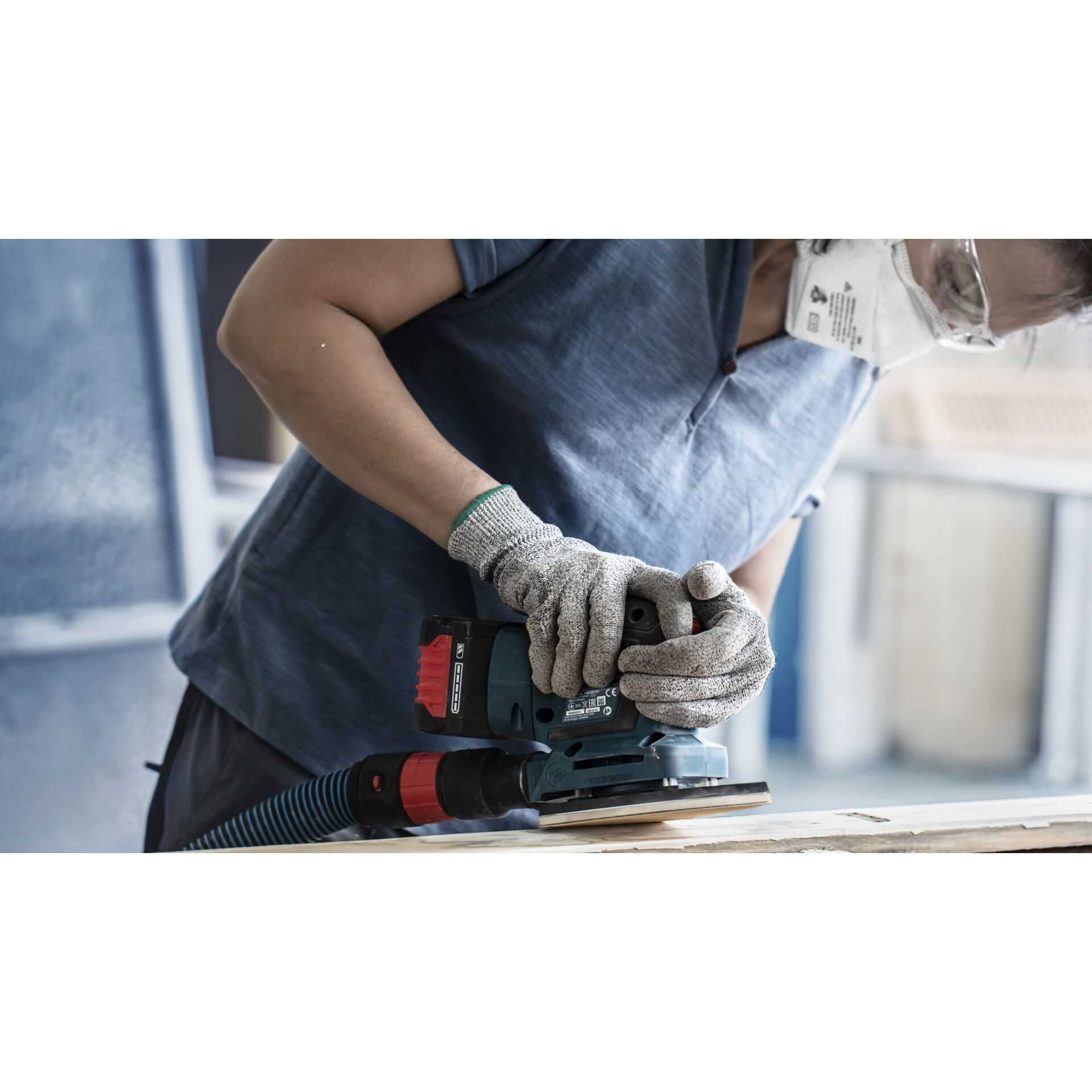 Eine Person schleift Holz mit einem Bosch-Schleifer, trägt Maske und Handschuhe. Blaues Hemd, graue Handschuhe.