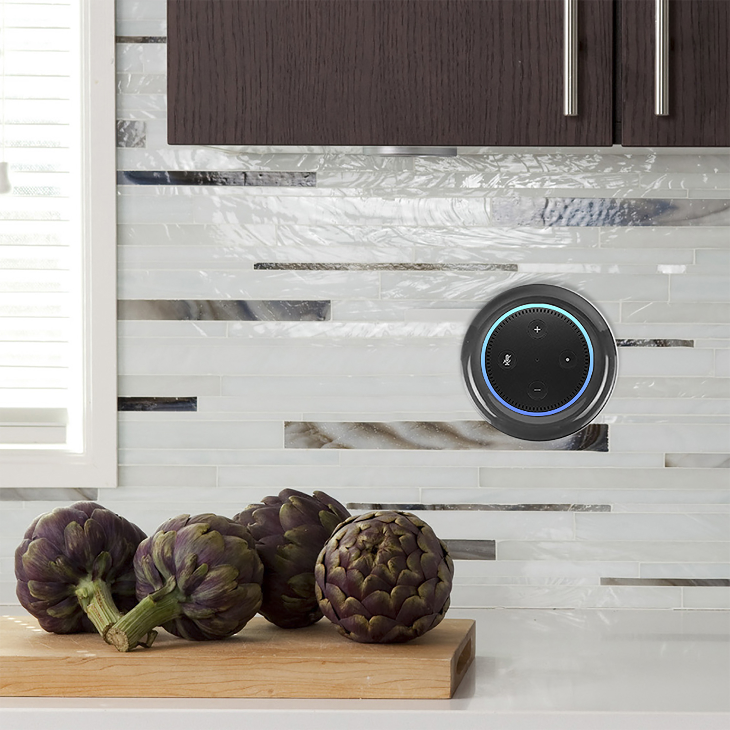 Un Echo Dot su una parete della cucina vicino a carciofi su un tagliere di legno con paraschizzi di piastrelle bianche.