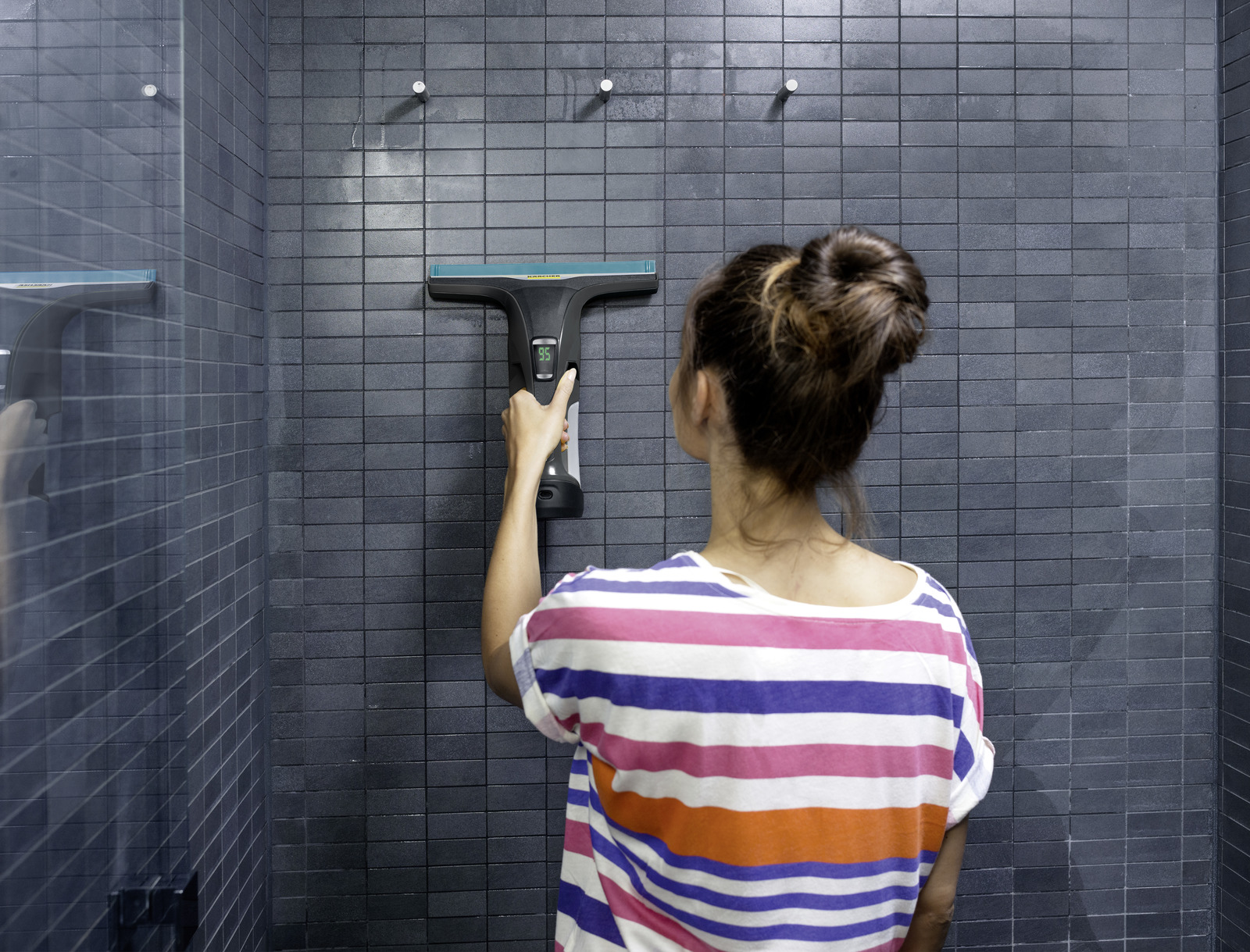 Vrouw maakt douchewand schoon met raamtrekker. Gestreepte shirt en knot.