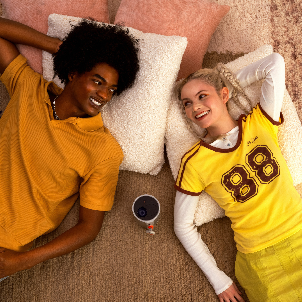 Para leży, uśmiechając się. Aparat i poduszki na łóżku. Patrzą na siebie.