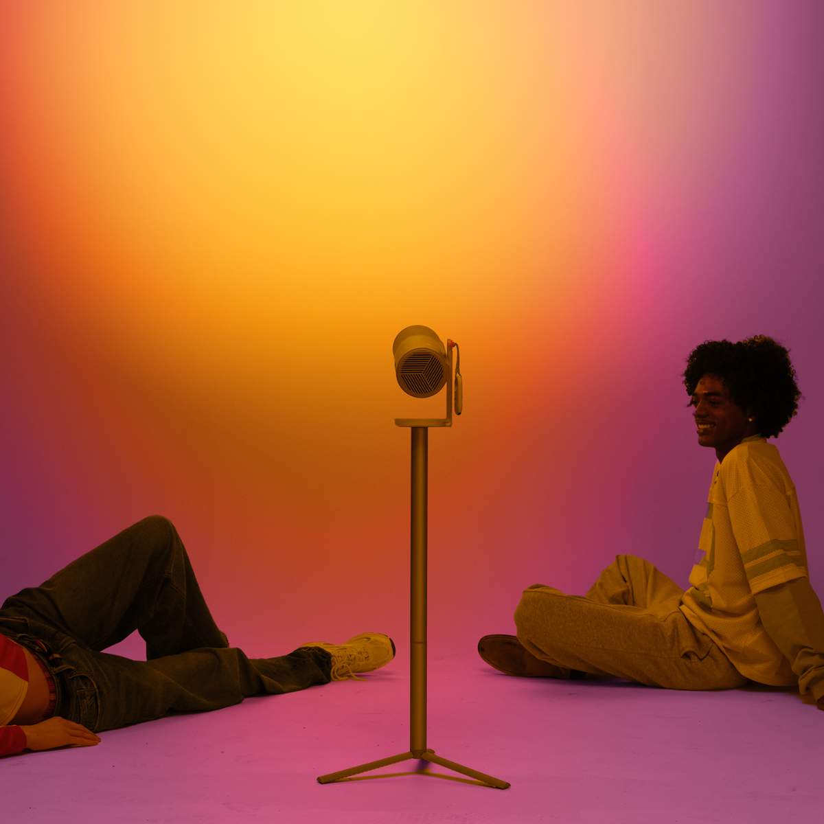 Dos personas tumbadas en un suelo rosa, iluminadas por una luz sobre un soporte con fondo degradado.