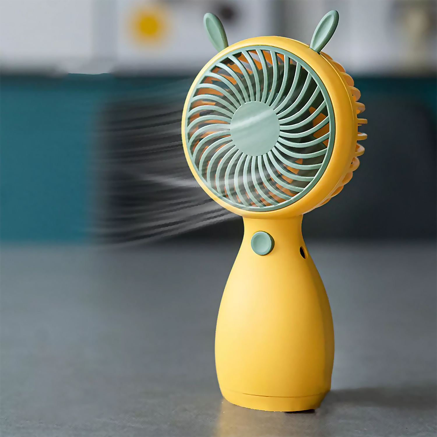 Ventilatore portatile giallo con dettagli verdi, aria che soffia. Superficie grigia di sfondo.