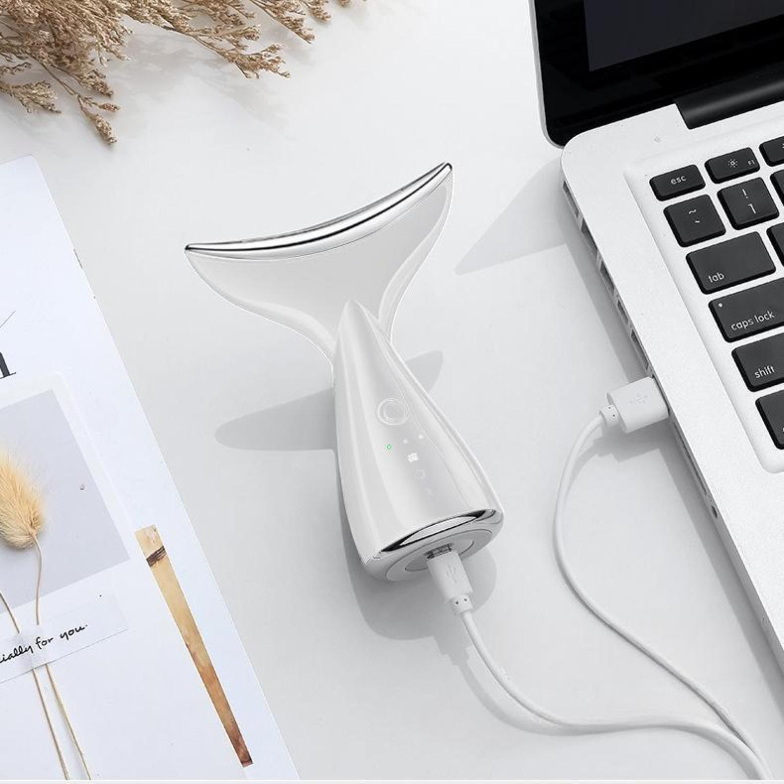 Weißes Gesichtsgerät mit USB-Ladekabel, auf einem weißen Tisch in der Nähe eines Laptops platziert.
