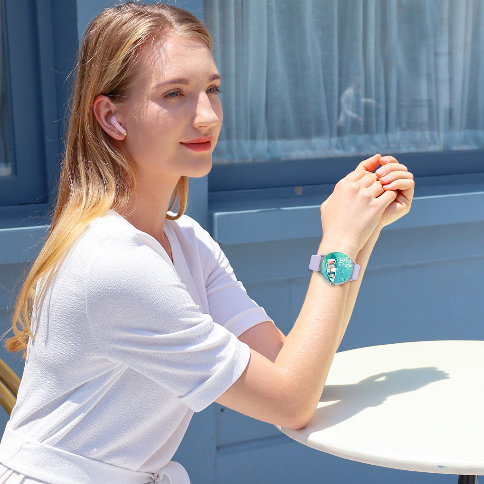 Frau mit Uhr und Ohrhörern sitzt an einem Tisch. Blaue Tür im Hintergrund.