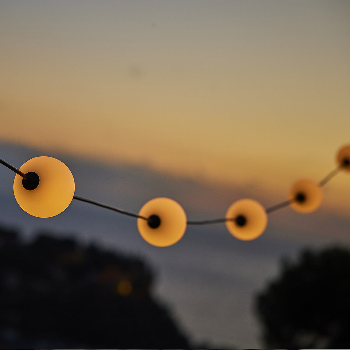Eine Lichterkette. Die Lichter sind weiße Kugeln. Der Hintergrund ist ein Sonnenuntergang.