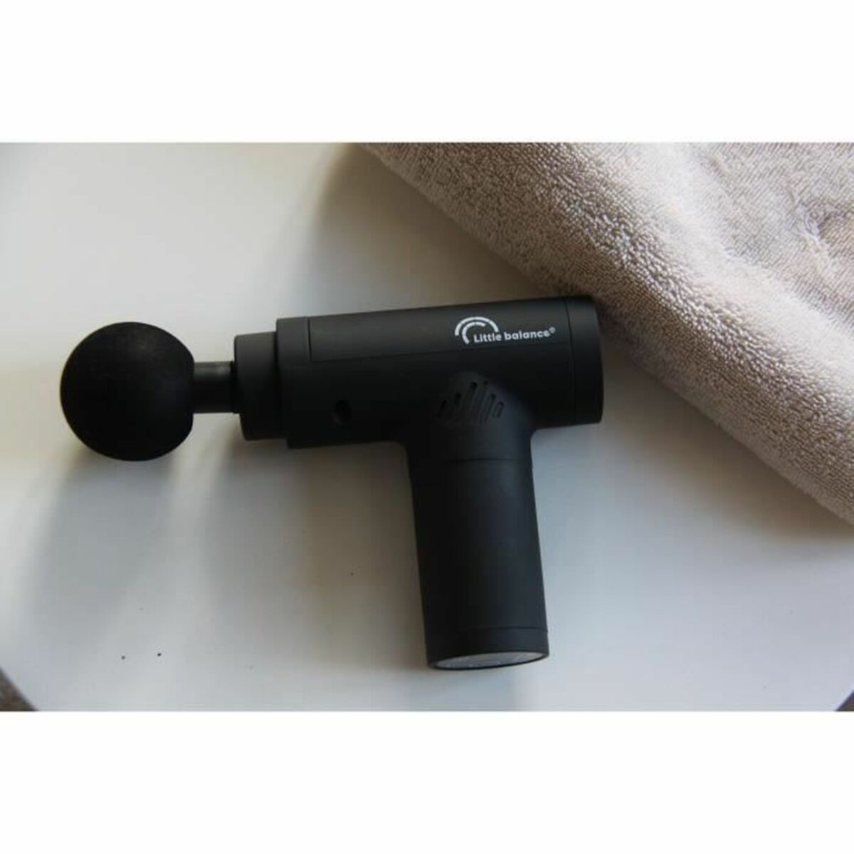 Schwarze Massagepistole auf weißer Oberfläche neben einem gefalteten grauen Handtuch.