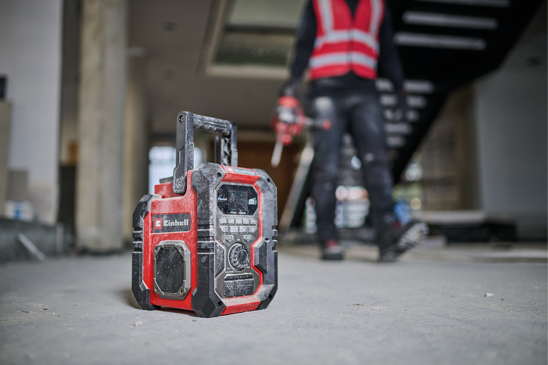 Ein rot-schwarzes Radio auf einer Baustelle mit einer Person im Hintergrund, die eine Weste trägt.