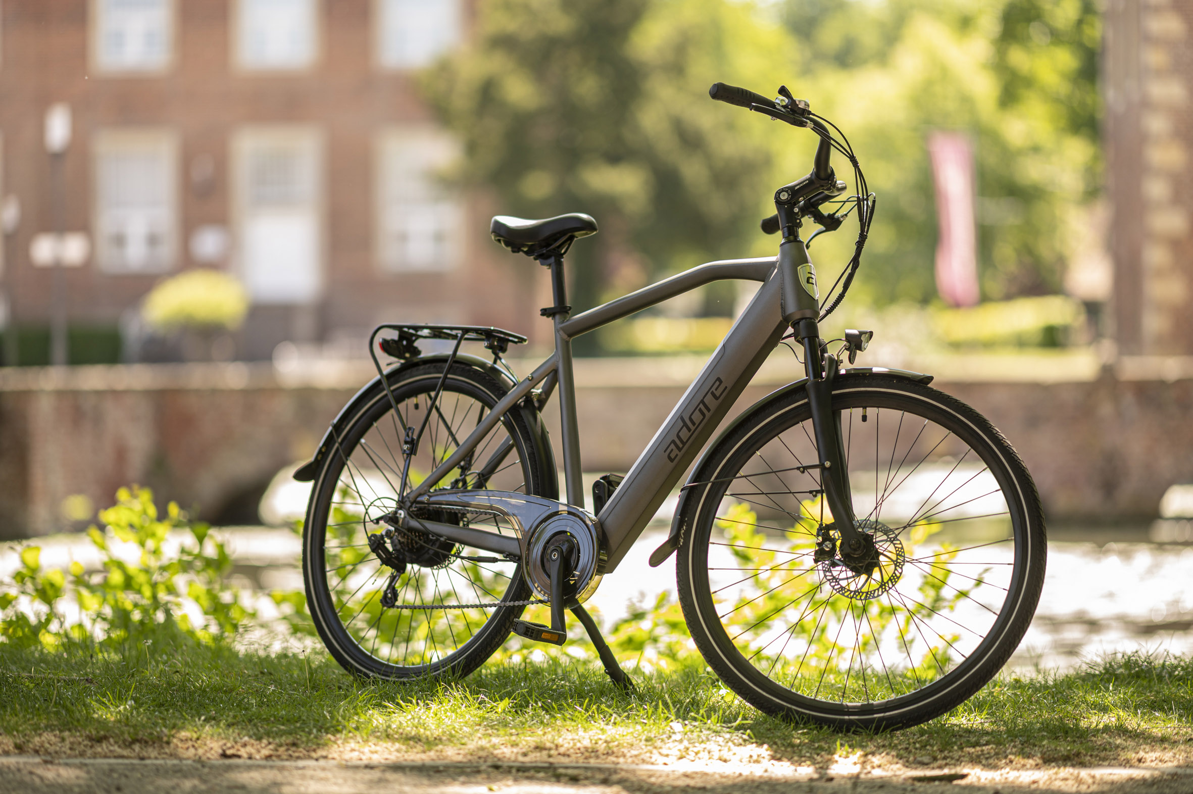 Ein graues Adore-Fahrrad auf Gras mit einem Backsteingebäude im Hintergrund.