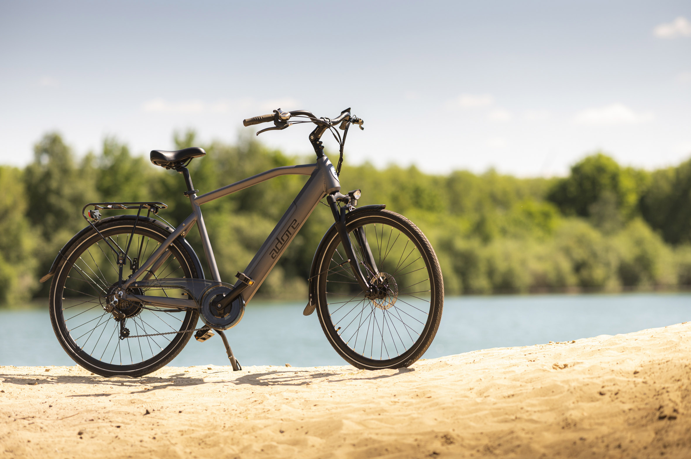 Ein graues Adore-Fahrrad auf sandigem Untergrund mit einem See und Bäumen im Hintergrund.