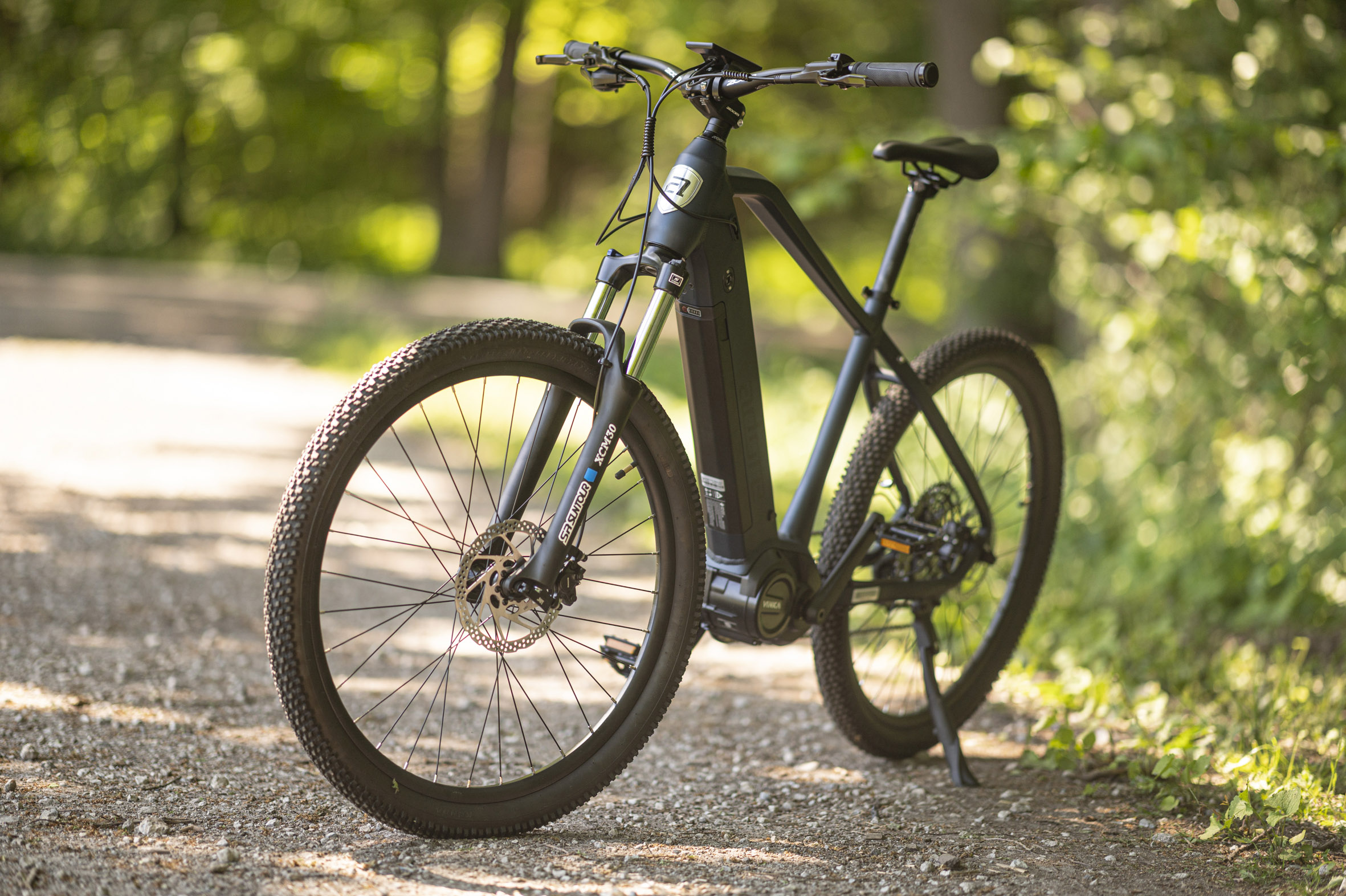 Ein schwarzes E-Bike auf einem Schotterweg. Grünes Laub umgibt es. Das Fahrrad hat Scheibenbremsen und Federung.