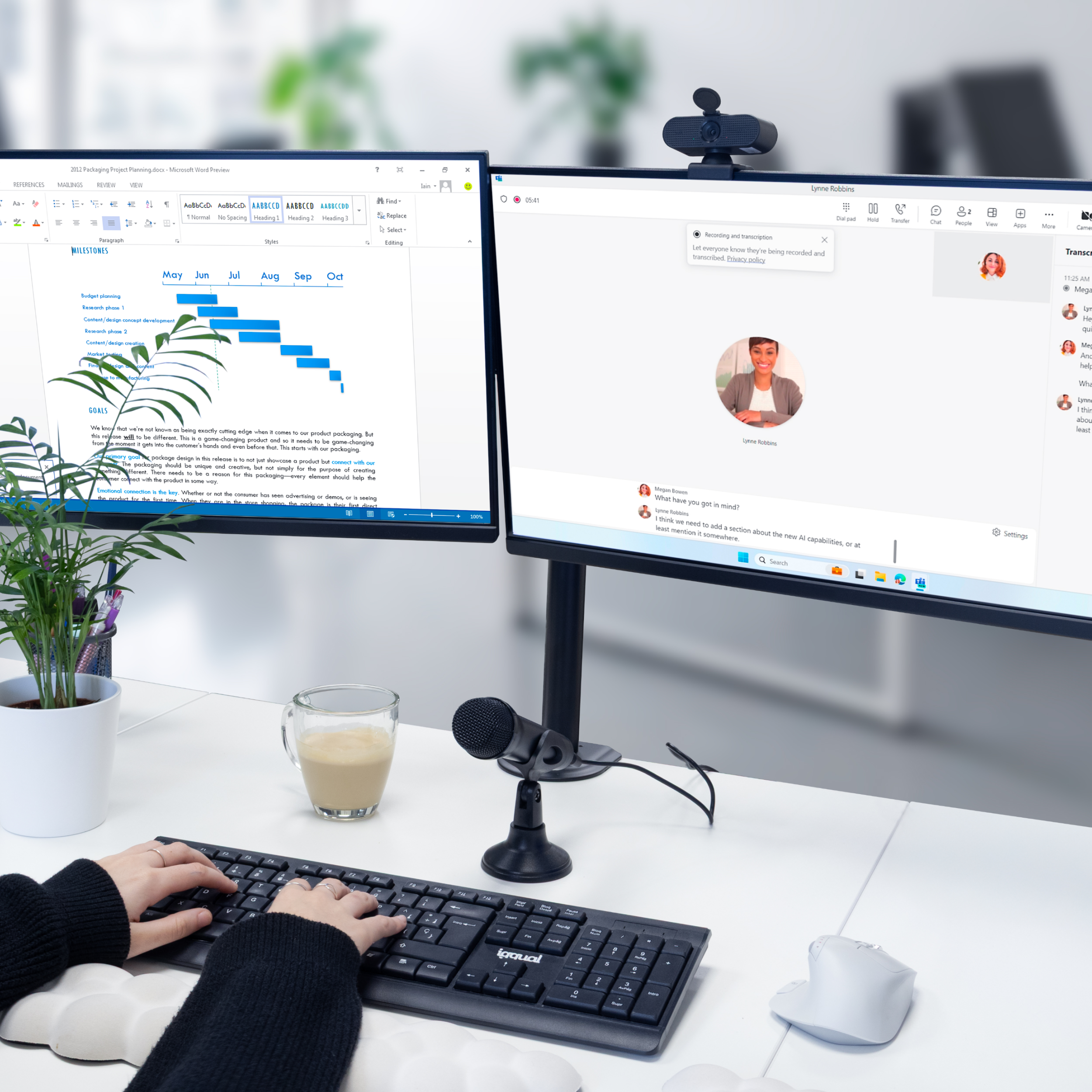 Osoba pisze na klawiaturze, dwa monitory z planami projektów i rozmową wideo. Filiżanka kawy i mikrofon.