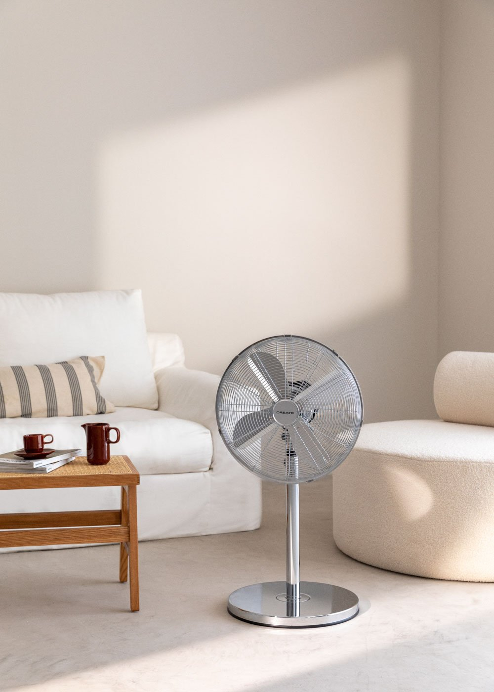 Un ventilador de pie en una habitación luminosa. Un sofá blanco y una mesa de centro en el fondo.