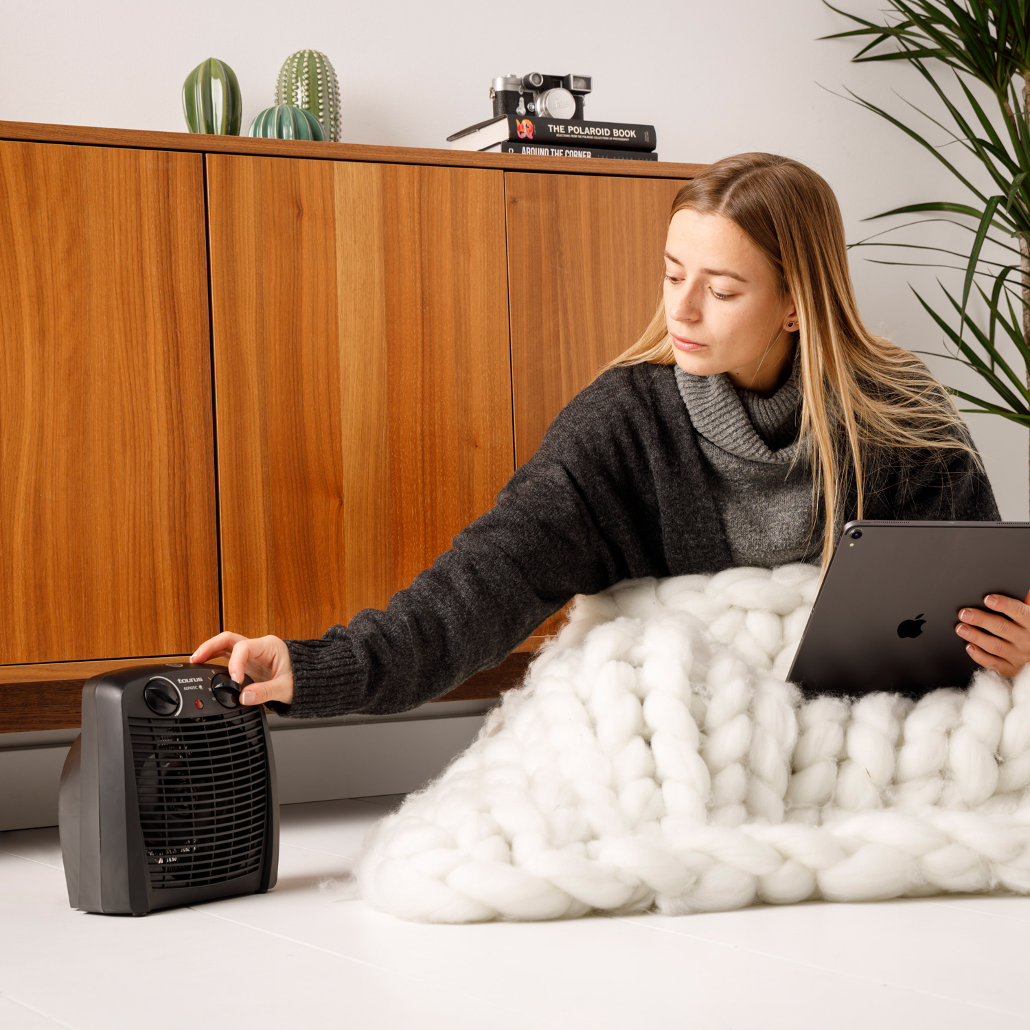 Kobieta siedzi, używa tabletu, reguluje grzejnik, obok koca i szafki.