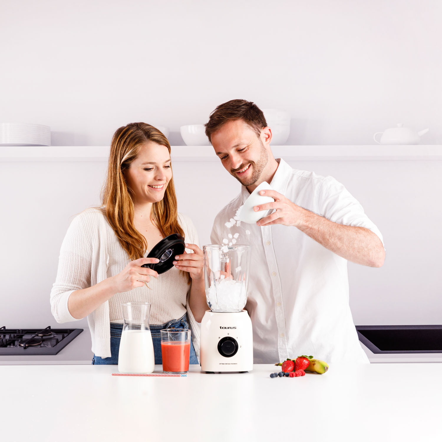 Ein Paar macht einen Smoothie mit einem Mixer, Früchten und Gläsern auf einer weißen Arbeitsplatte.