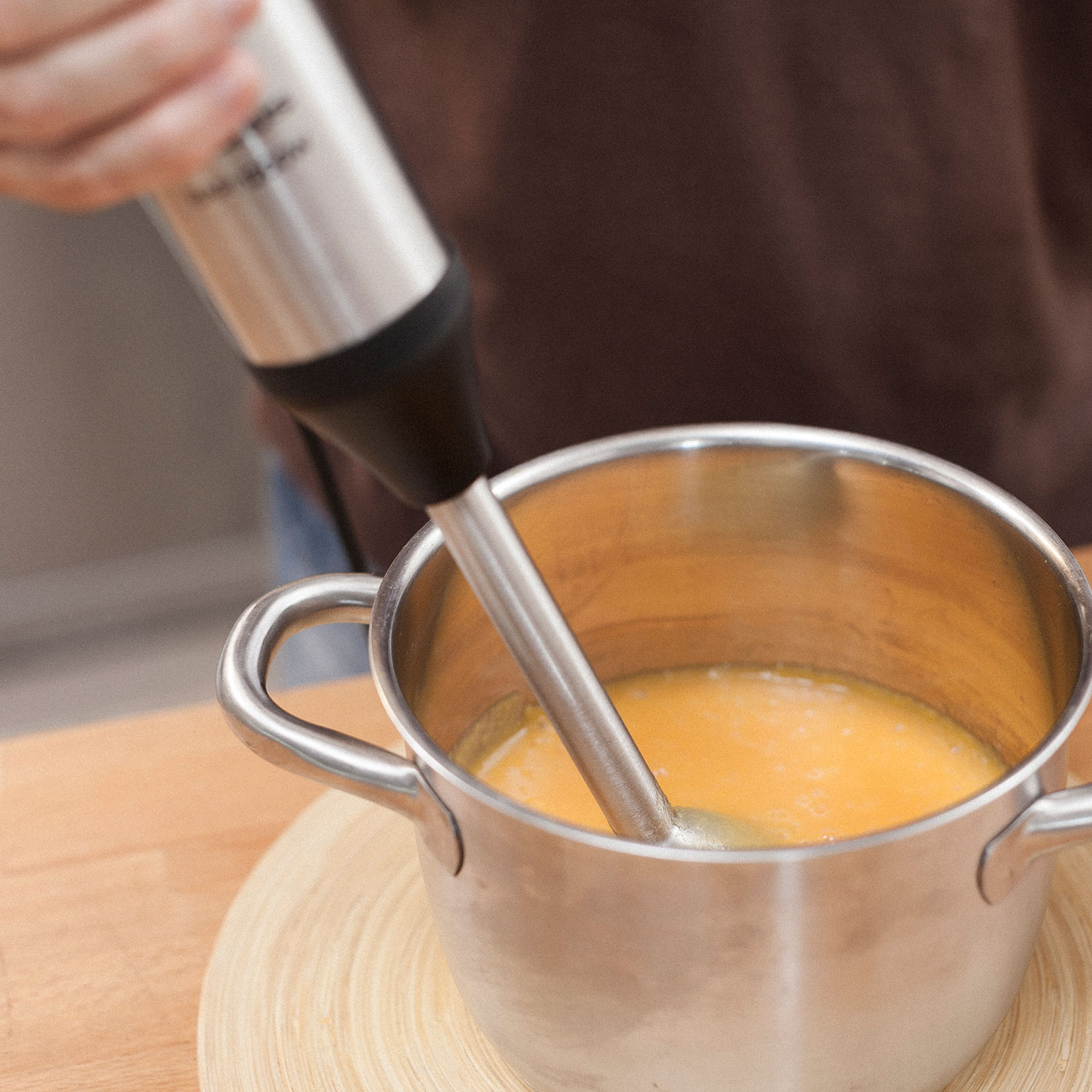 Ręka trzymająca blender ręczny w garnku z pomarańczową zupą, na drewnianej powierzchni.