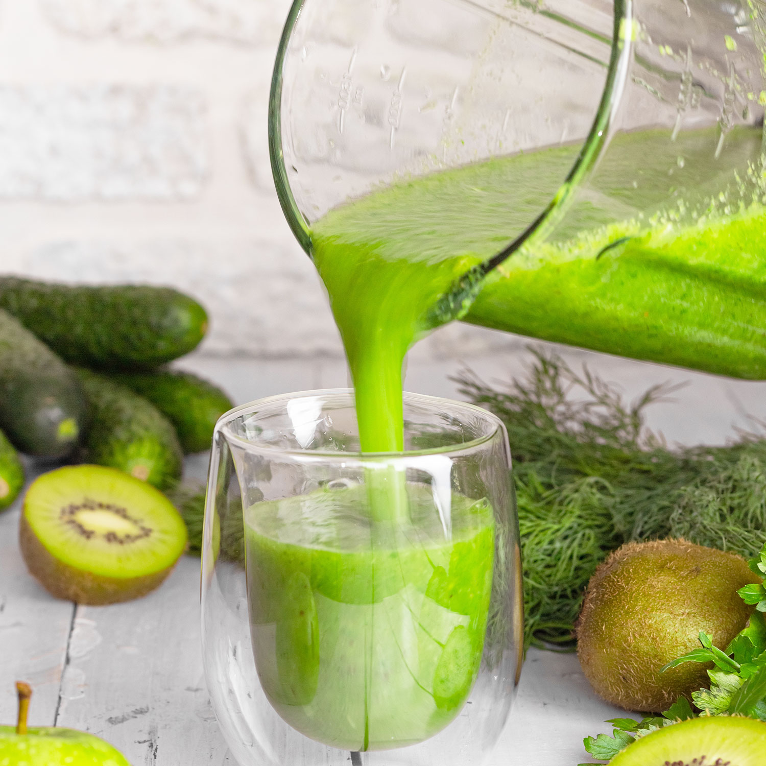 Un batido verde se vierte de una licuadora a un vaso. Kiwis y pepinos están cerca.
