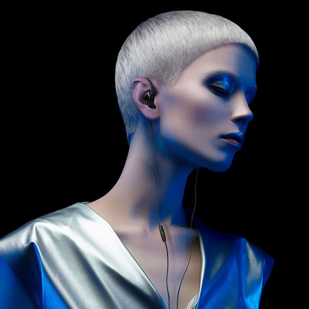 Frau mit kurzen weißen Haaren trägt silbernes Oberteil, Ohrhörer, blaues Licht, schwarzer Hintergrund.