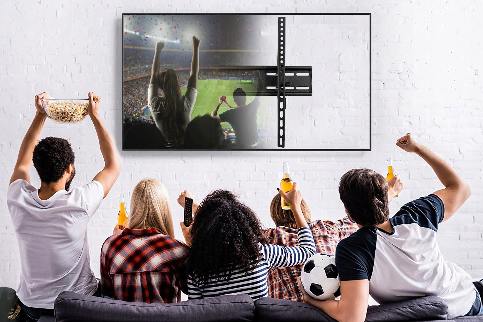 Ludzie oglądają telewizję. Niektórzy kibicują i trzymają napoje i przekąski. Duży telewizor na ścianie.