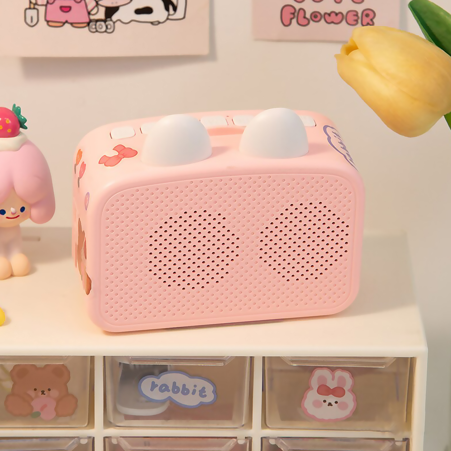 Un despertador rosa con orejas blancas y orificios para el altavoz está sobre un cajón. Una figura y un tulipán están en el fondo.