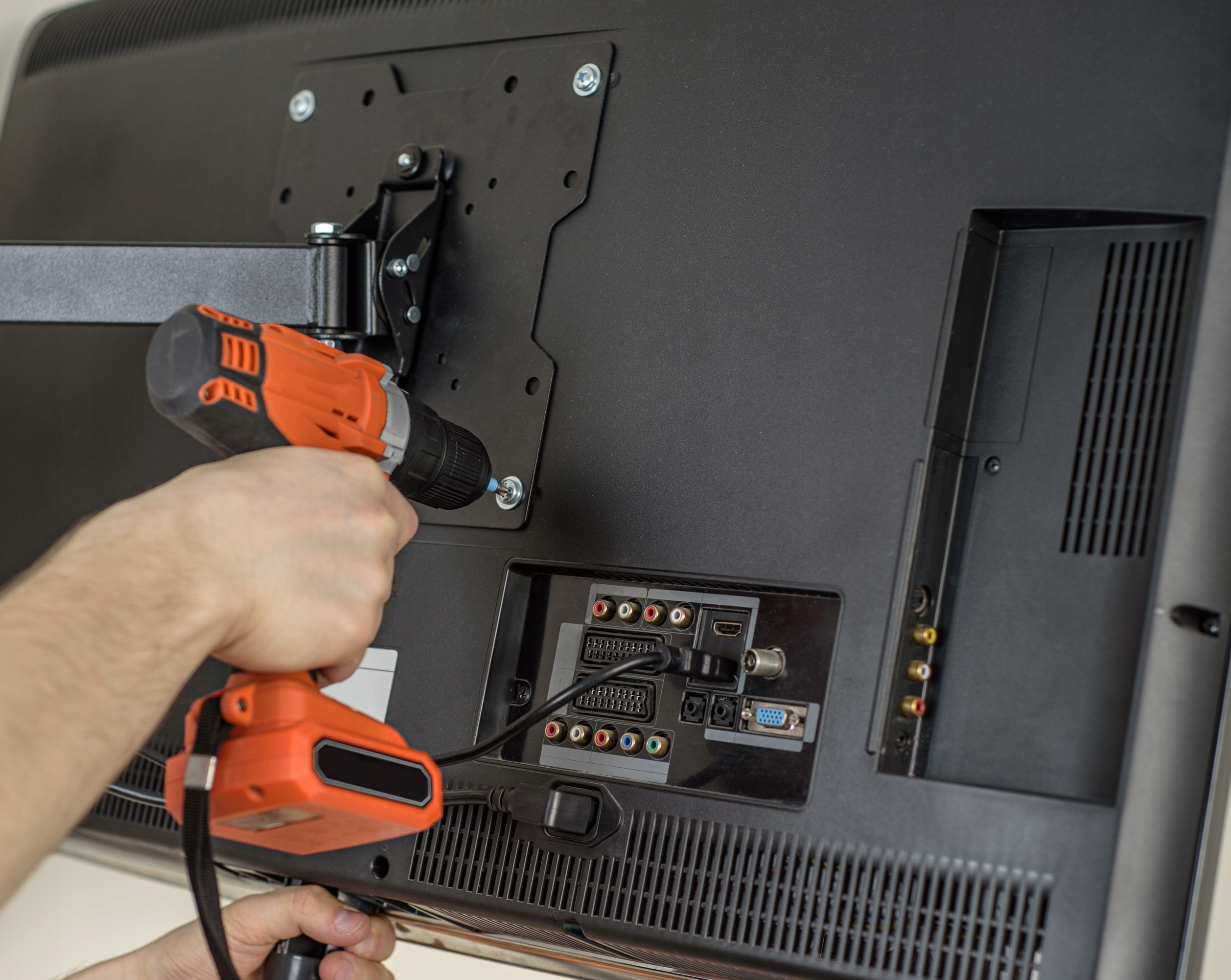 Osoba instaluje uchwyt do telewizora z tyłu telewizora za pomocą wiertarki. Pokazano zbliżenie tego procesu.