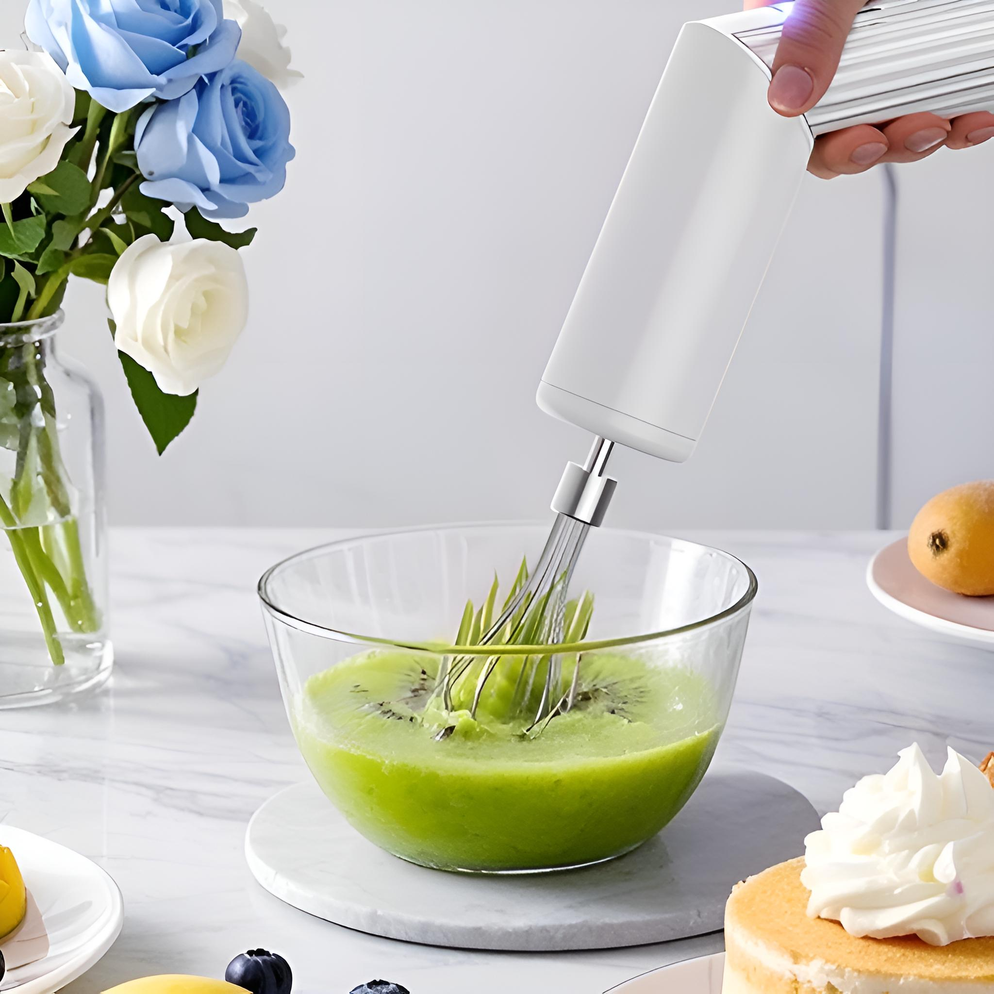Elektrischer Handmixer, der grüne Mischung in einer Glasschüssel verrührt. Kuchen und Obst sind sichtbar.