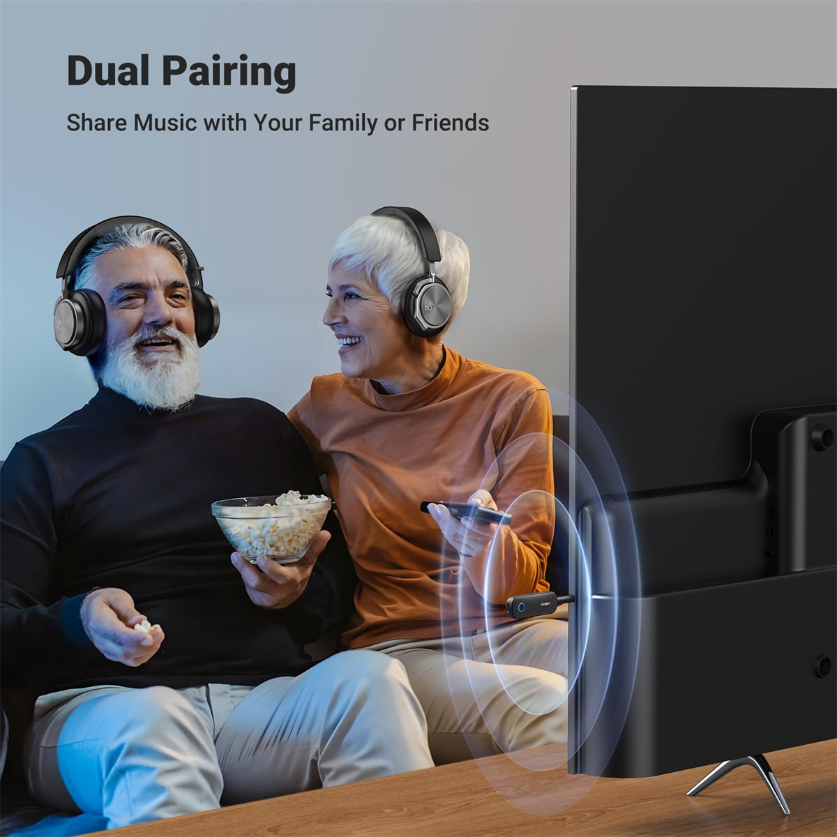 Älteres Ehepaar mit Kopfhörern, sitzt am Fernseher, einer hält Popcorn.