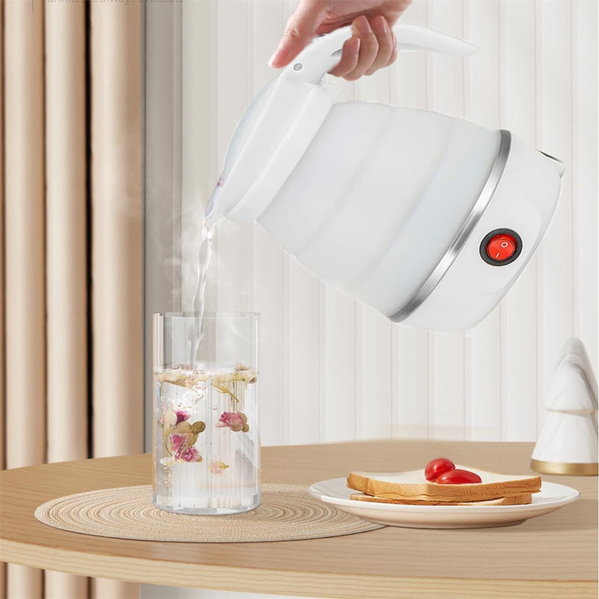 Ein weißer, zusammenklappbarer Wasserkocher gießt Wasser in ein klares Glas mit Blumen und eine Scheibe Brot mit Tomaten.