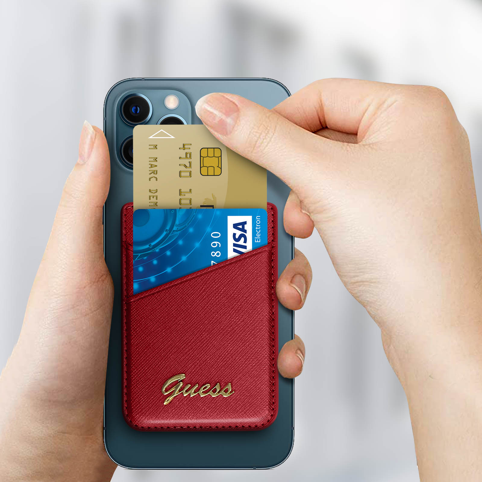 Mani che tengono una carta di credito blu e un portafoglio per telefono Guess rosso. Telefono sullo sfondo.