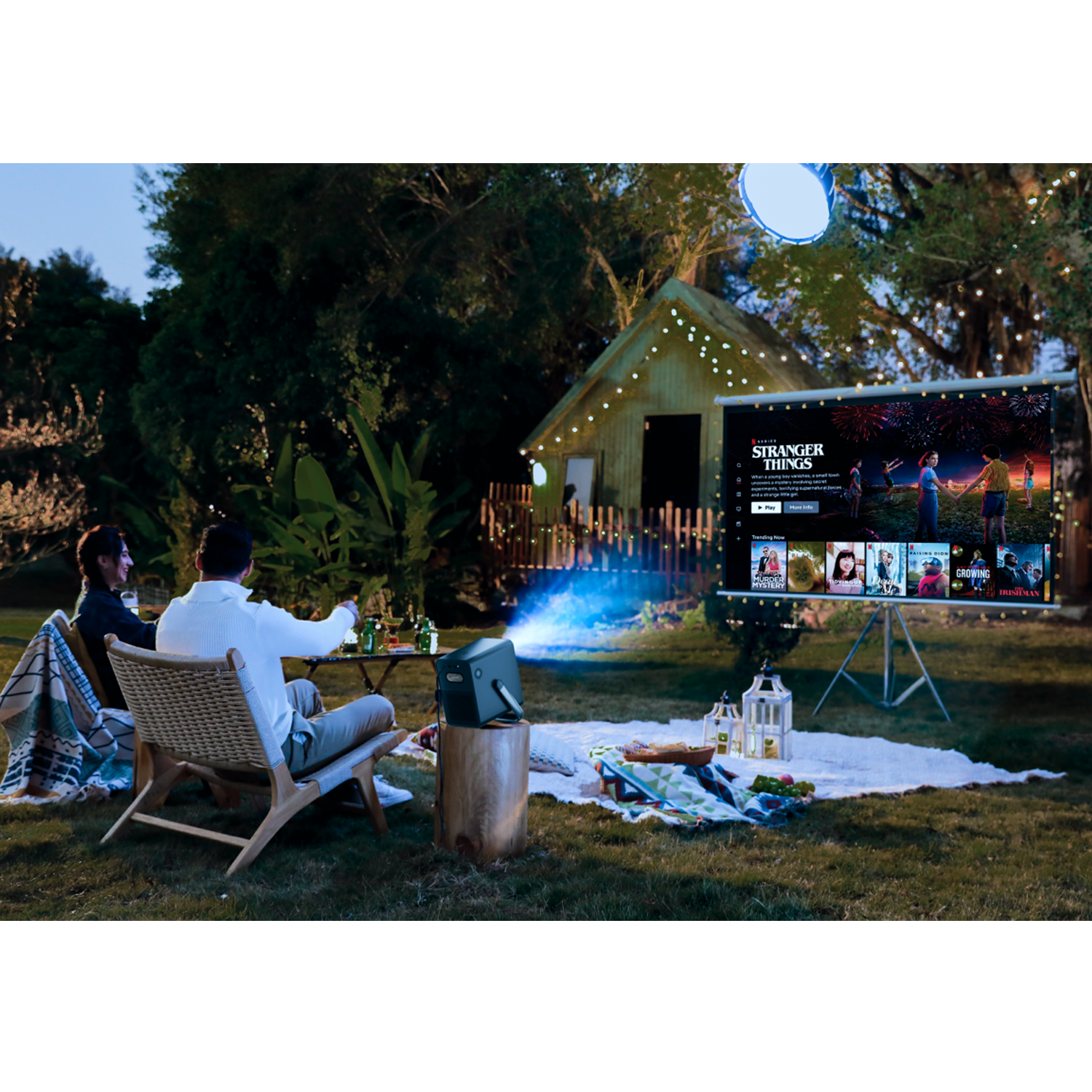 Mensen kijken buiten naar een film. Een projector op een tafel projecteert op een scherm. Stranger Things wordt getoond.