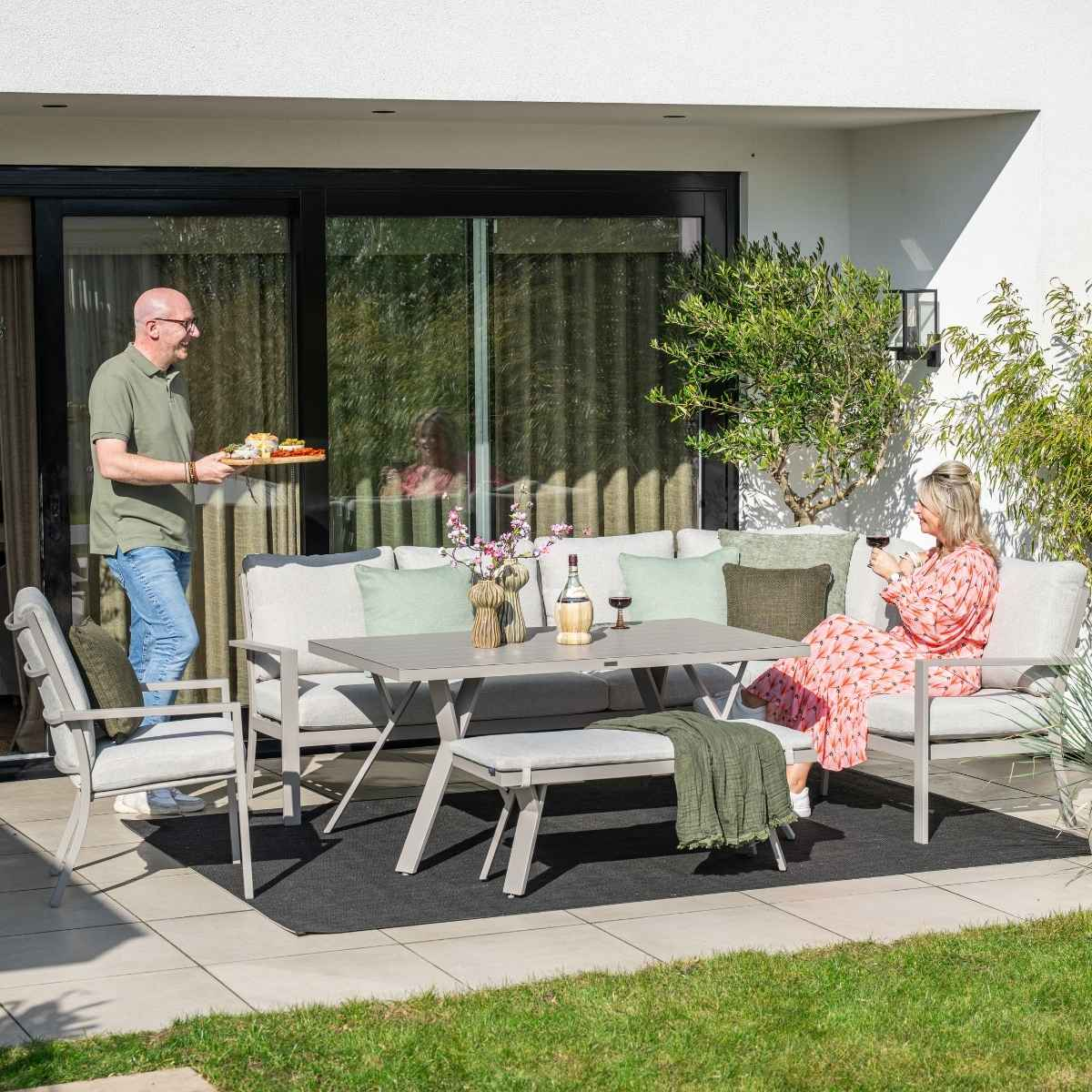 Homme servant de la nourriture, femme sur canapé avec du vin. Patio extérieur avec table, chaises et verdure.