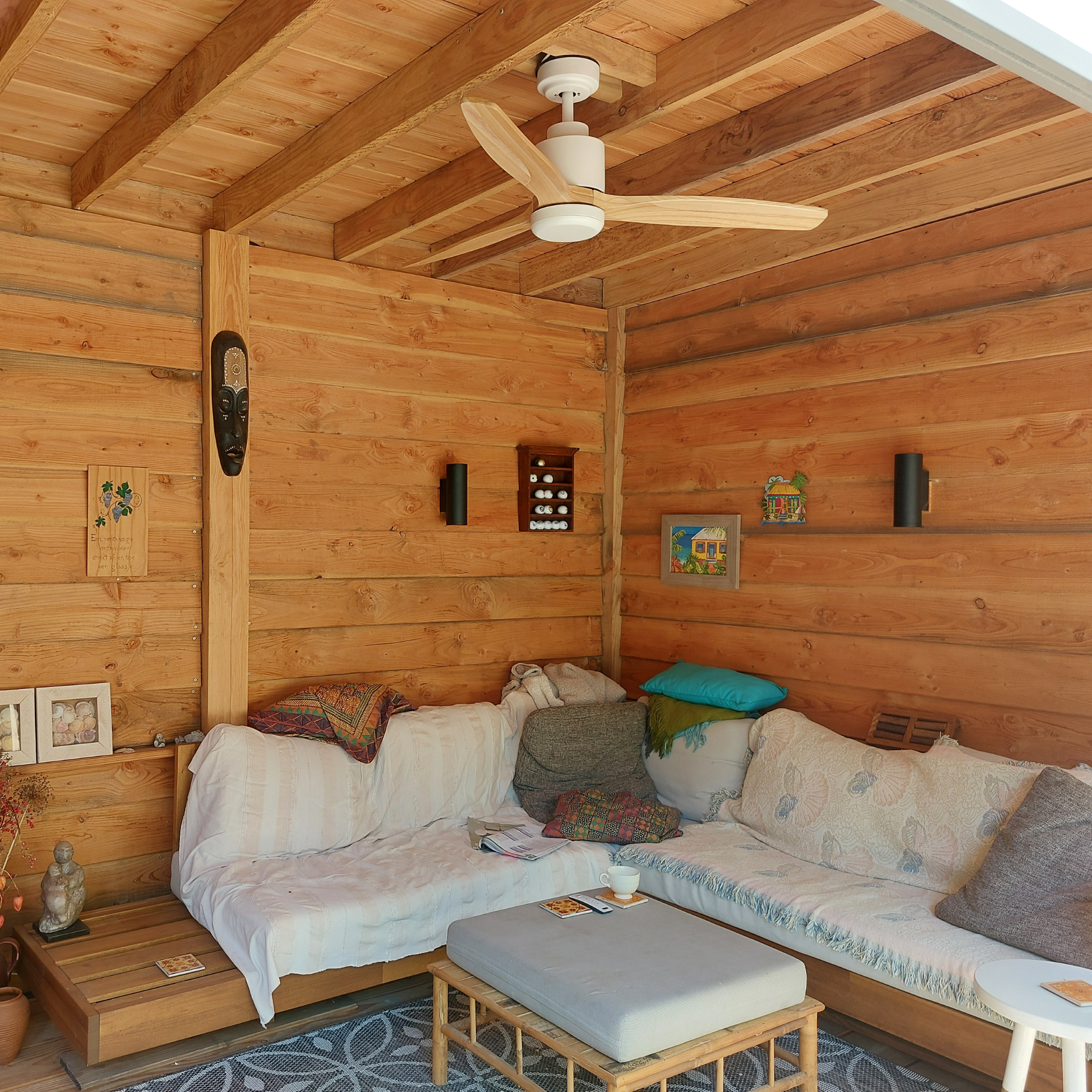 Interno di una cabina di legno con divani, cuscini e ventilatore a soffitto. Le decorazioni includono maschere e arte.