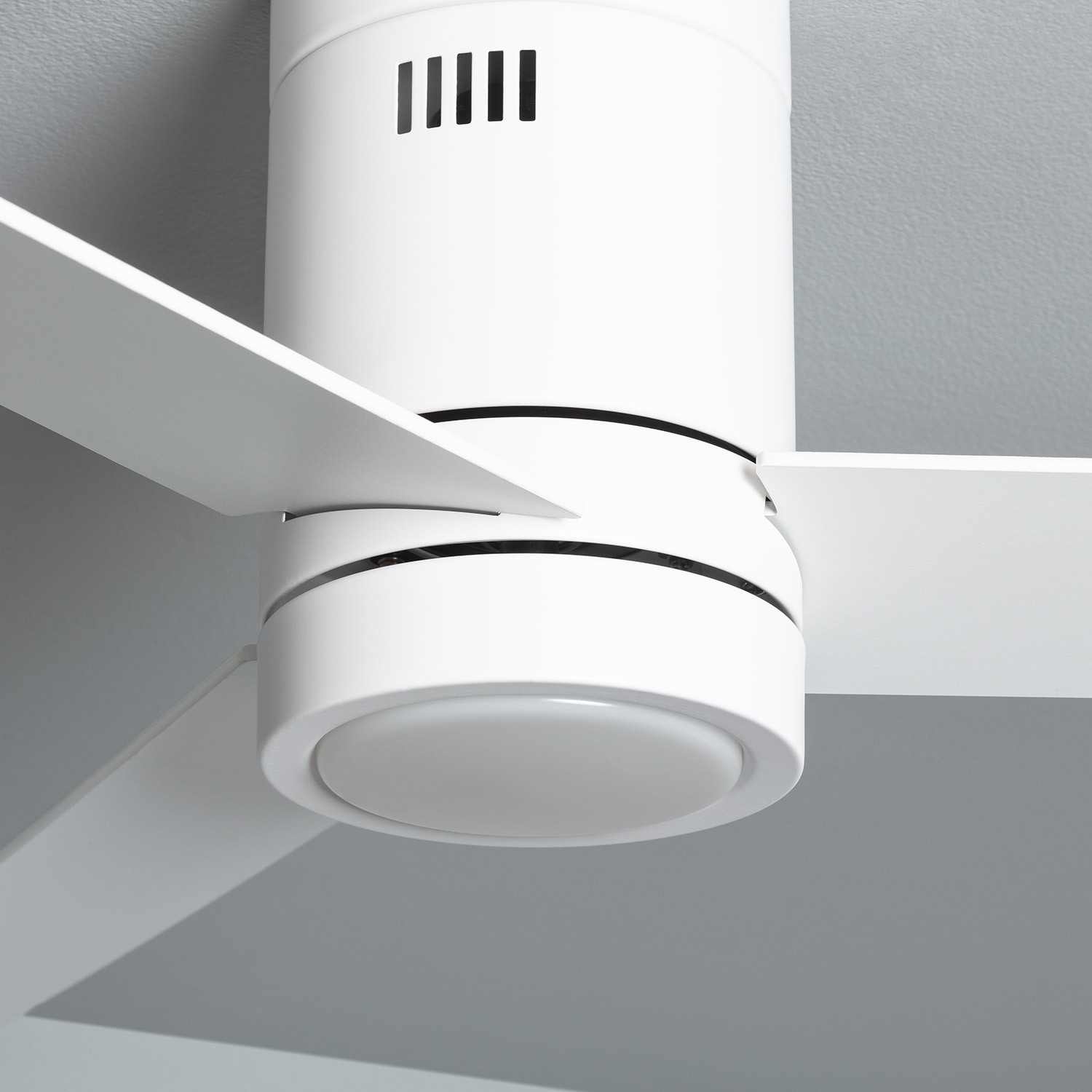 Primo piano di un ventilatore da soffitto bianco. Il ventilatore ha tre pale. Lo sfondo è grigio.