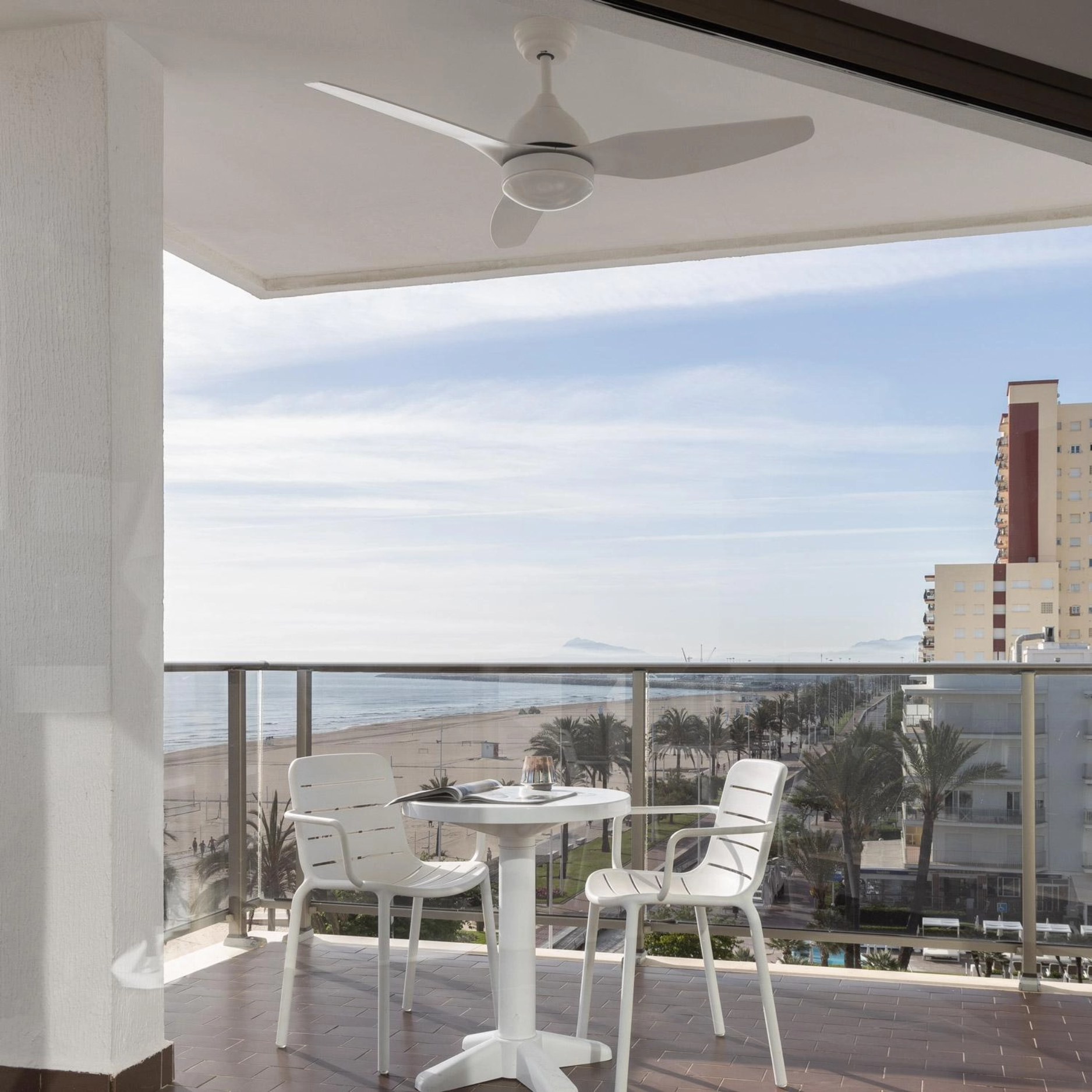 Balcone con tavolo e sedie bianche, con vista sulla spiaggia e sul mare. Ventilatore a soffitto ed edifici sullo sfondo.