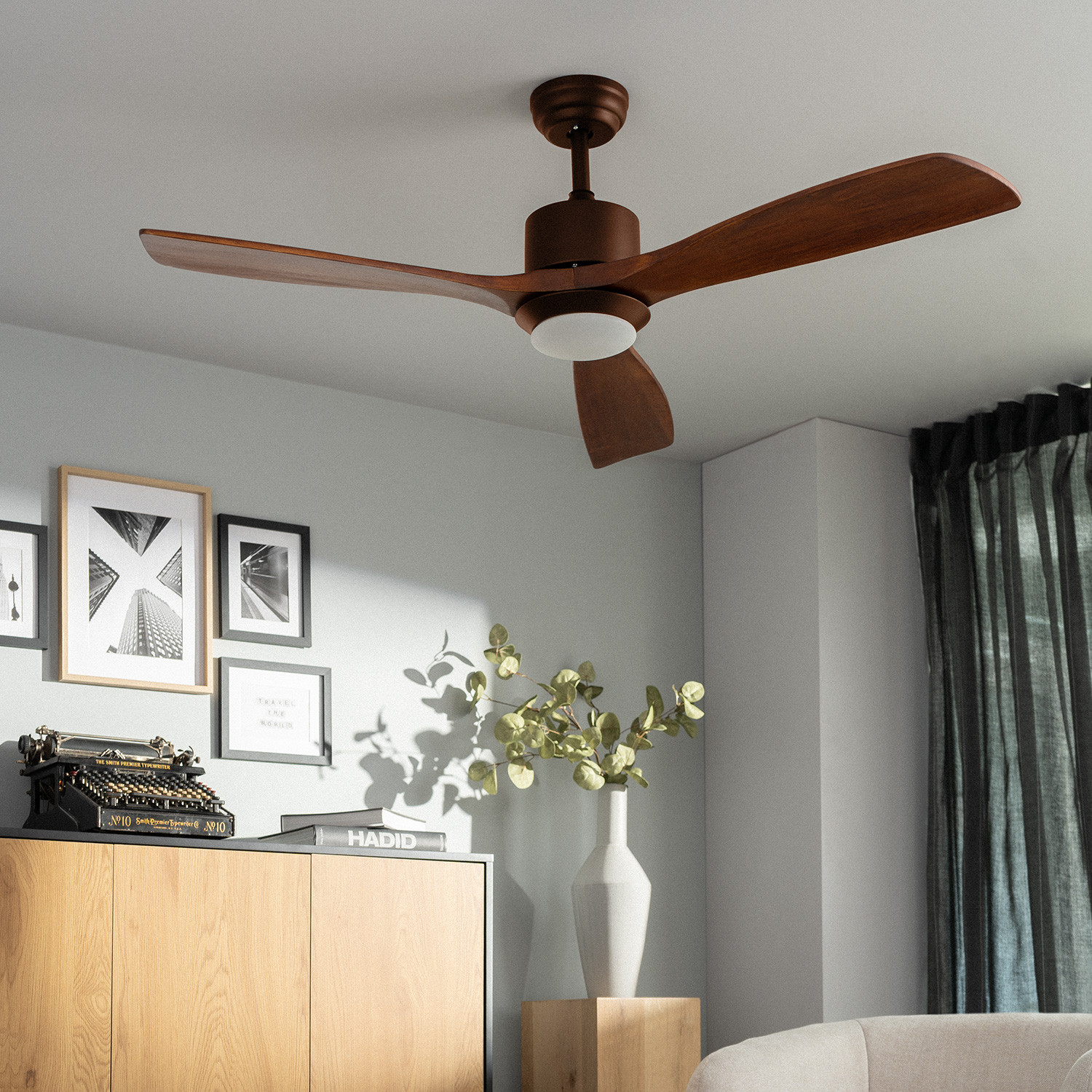 Un ventilatore a soffitto con tre pale in legno e una luce. La stanza ha arte e colori neutri.