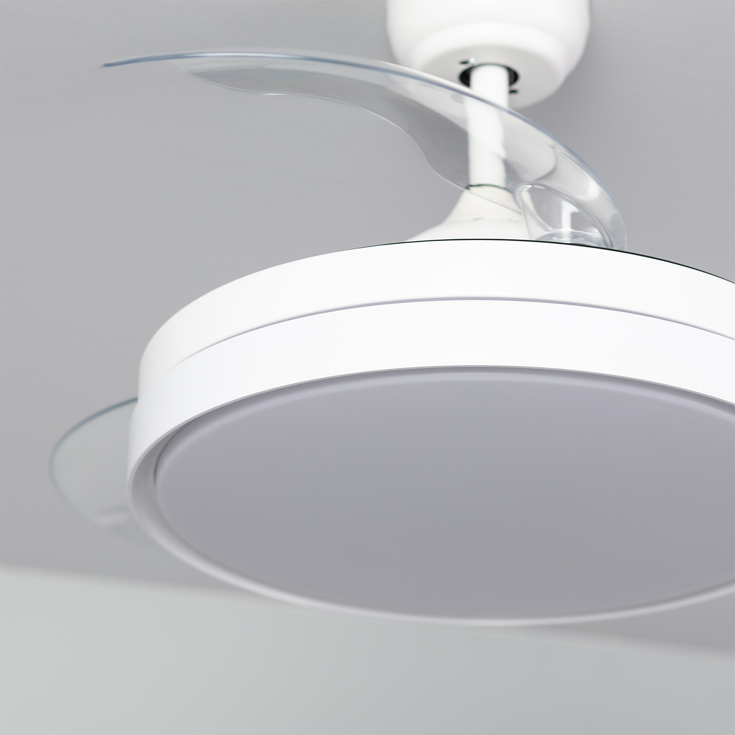 Primo piano di un ventilatore da soffitto bianco con pale trasparenti e un apparecchio di illuminazione circolare. Soffitto grigio sullo sfondo.