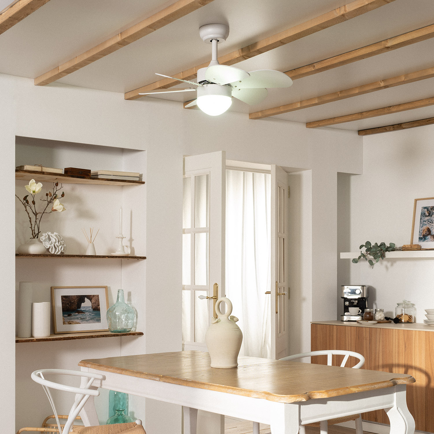 Sala da pranzo con pareti bianche, travi del soffitto in legno, tavolo, sedie, ventilatore a soffitto e oggetti decorativi.