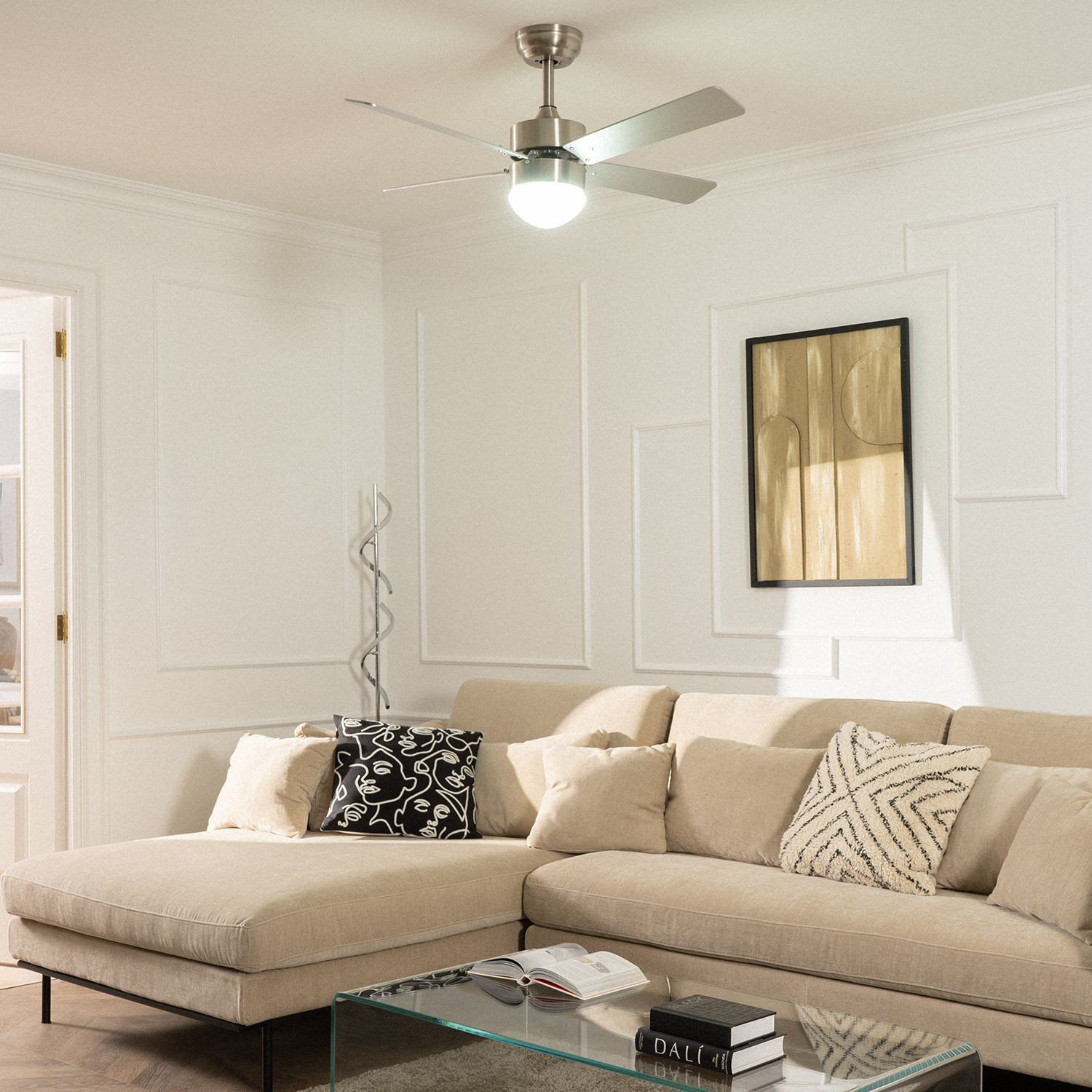 Soggiorno con divano beige, tavolo in vetro, ventilatore a soffitto. Arte sul muro, porta aperta, pareti bianche.