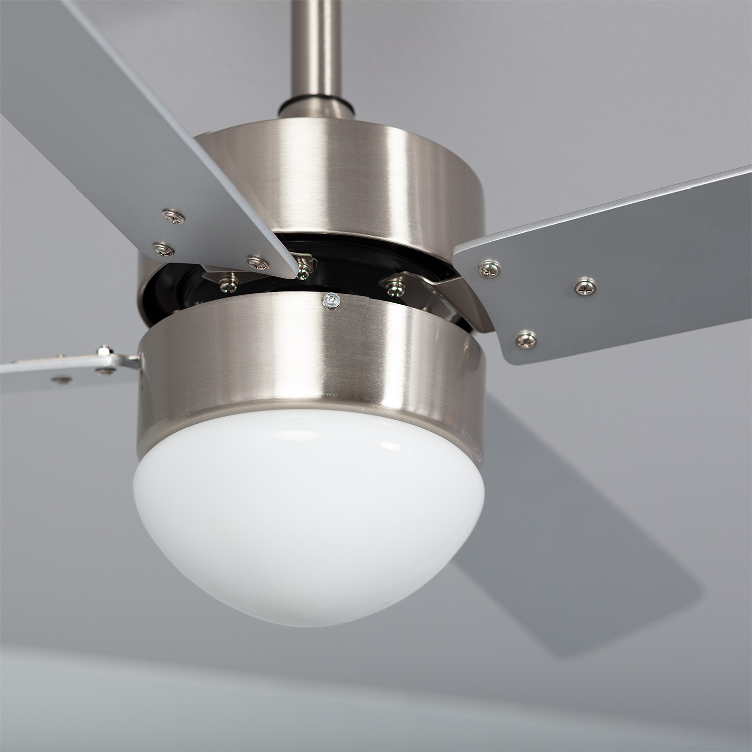 Primo piano del ventilatore a soffitto. Pale argentate, lampada bianca. Soffitto bianco. Ombra visibile.