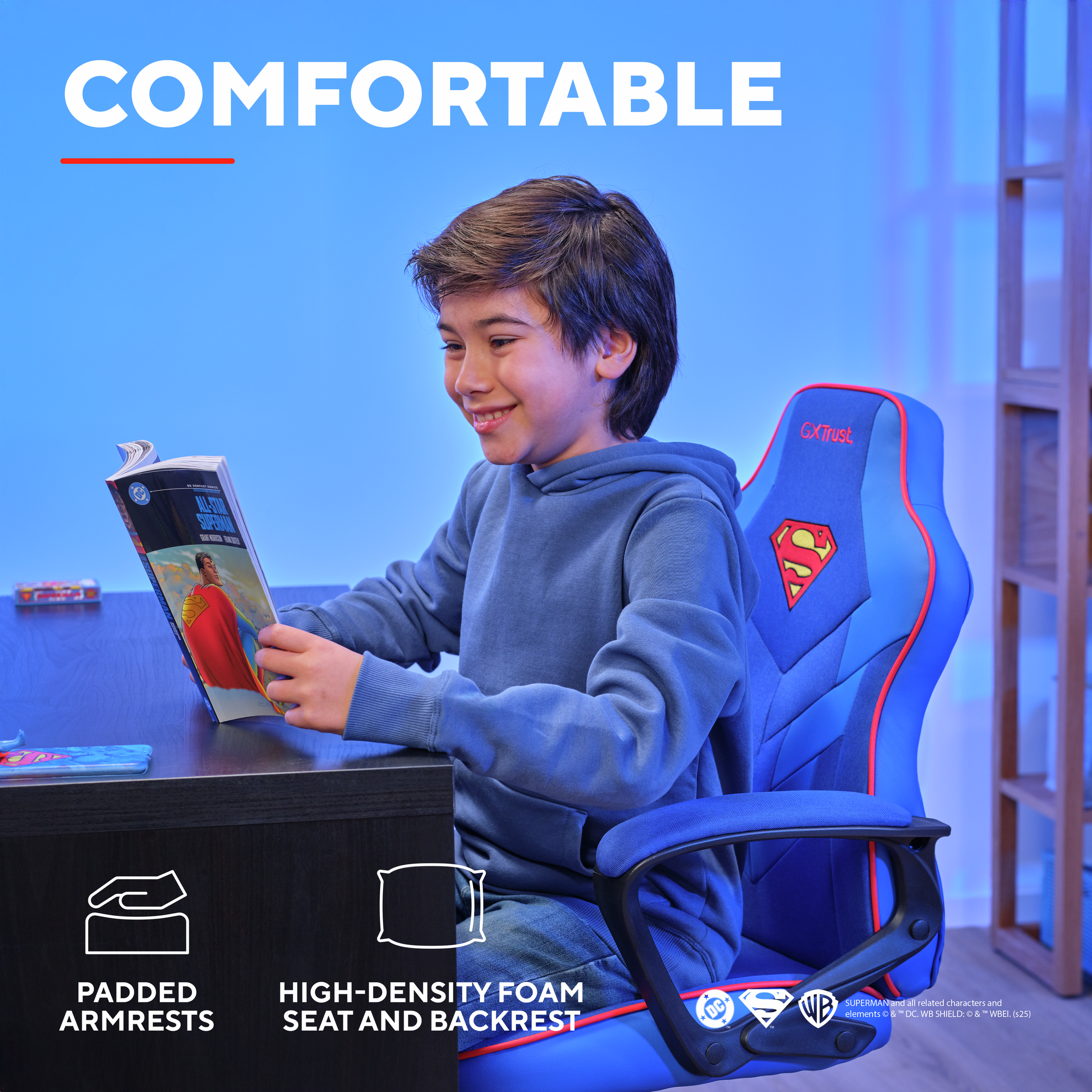 Chłopiec czytający komiks o Supermanie, siedzący na niebieskim krześle Supermana. Czarne biurko i tło.