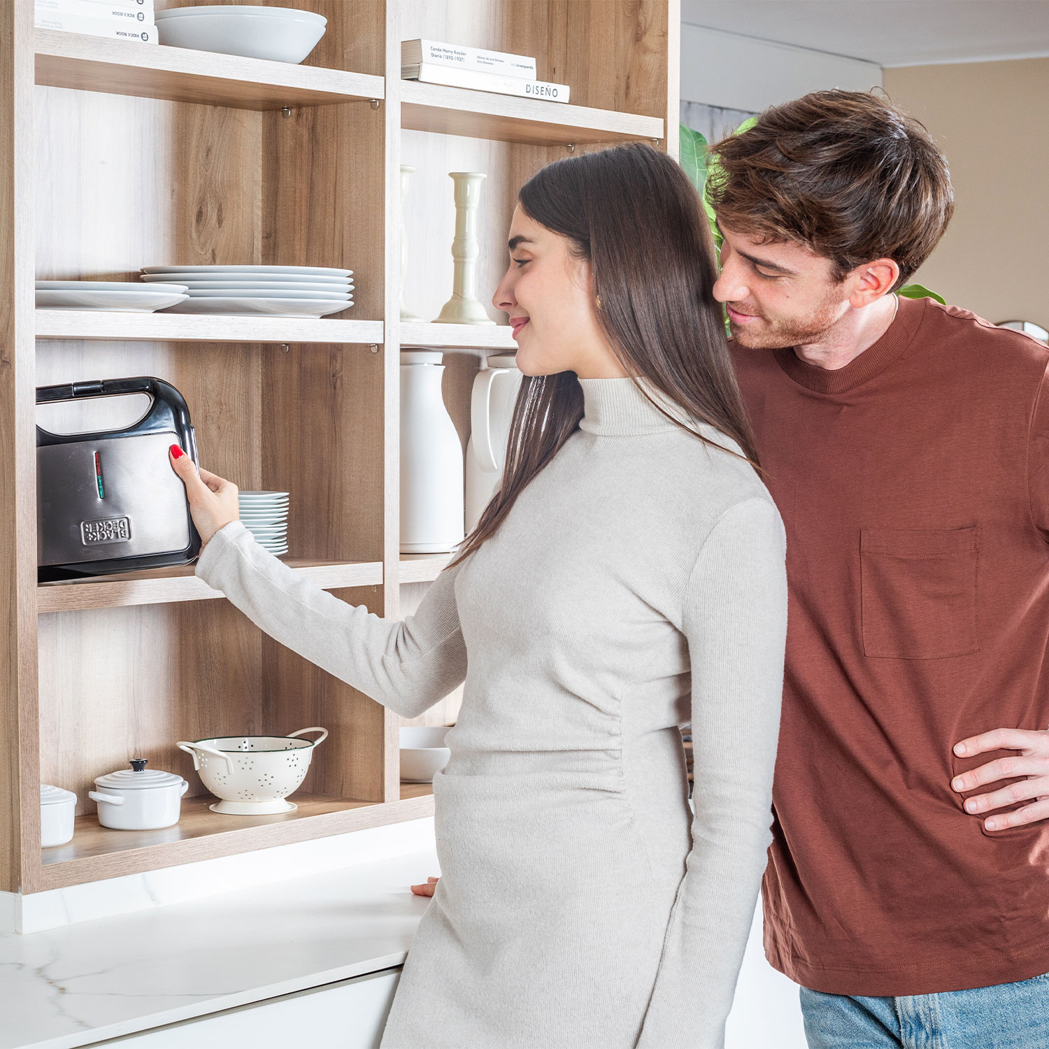 Para w kuchni patrzy na czarne urządzenie na półce, kobieta wskazuje na nie.