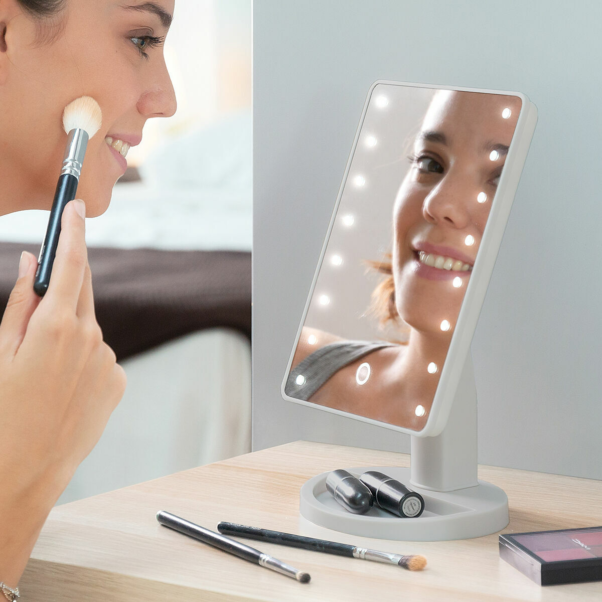 Frau, die Make-up mit einem Pinsel und einem beleuchteten Spiegel aufträgt. Lippenstift und Pinsel auf dem Tisch.